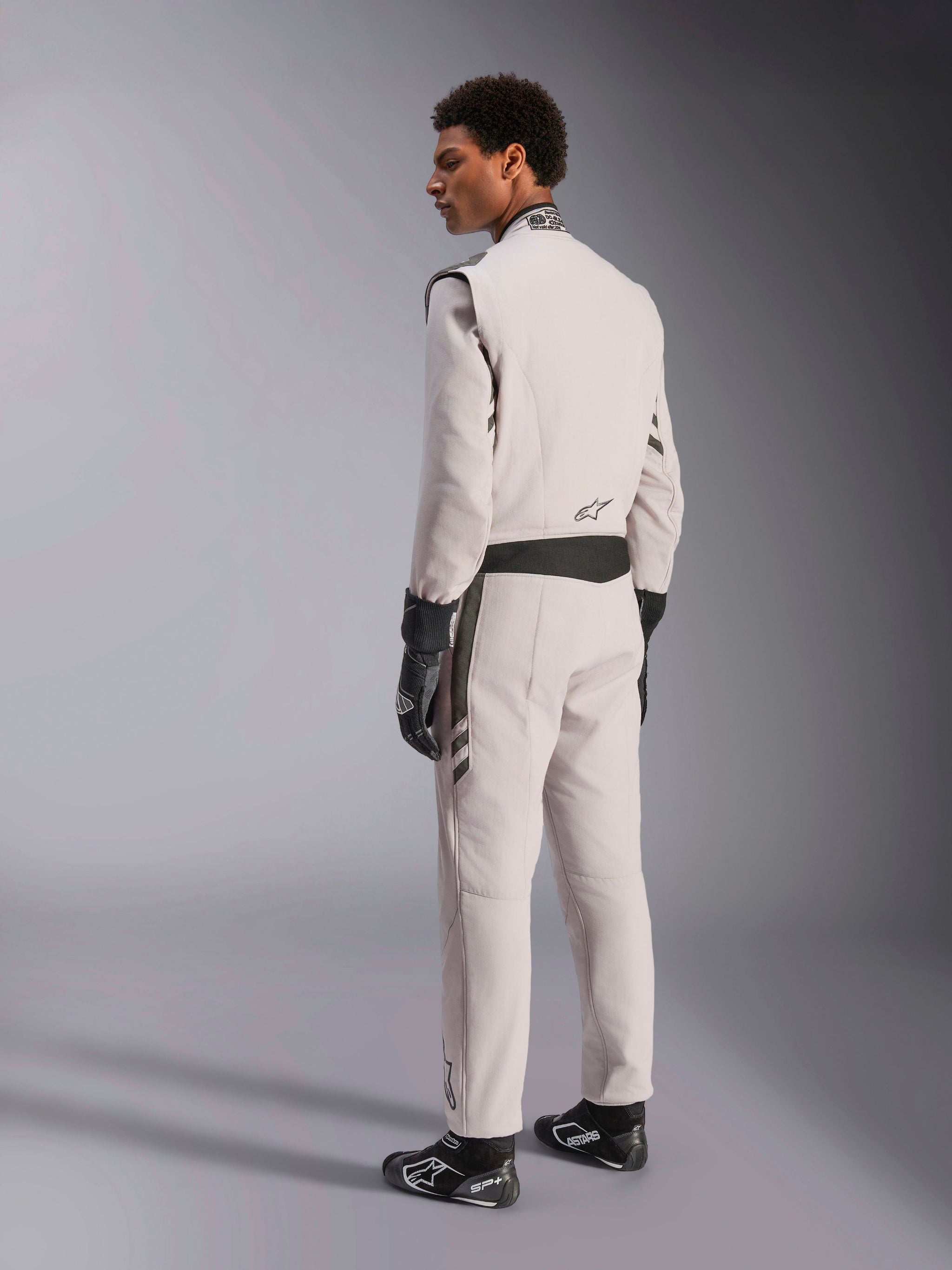 Tuta Alpinestars GP Race V3 FIA, tuta da corsa Silver Anthracite, vista laterale e posteriore, indossata con guanti e stivali da corsa neri, con accenti grigio scuro su vita e spalle, abbigliamento protettivo professionale per automobilismo.