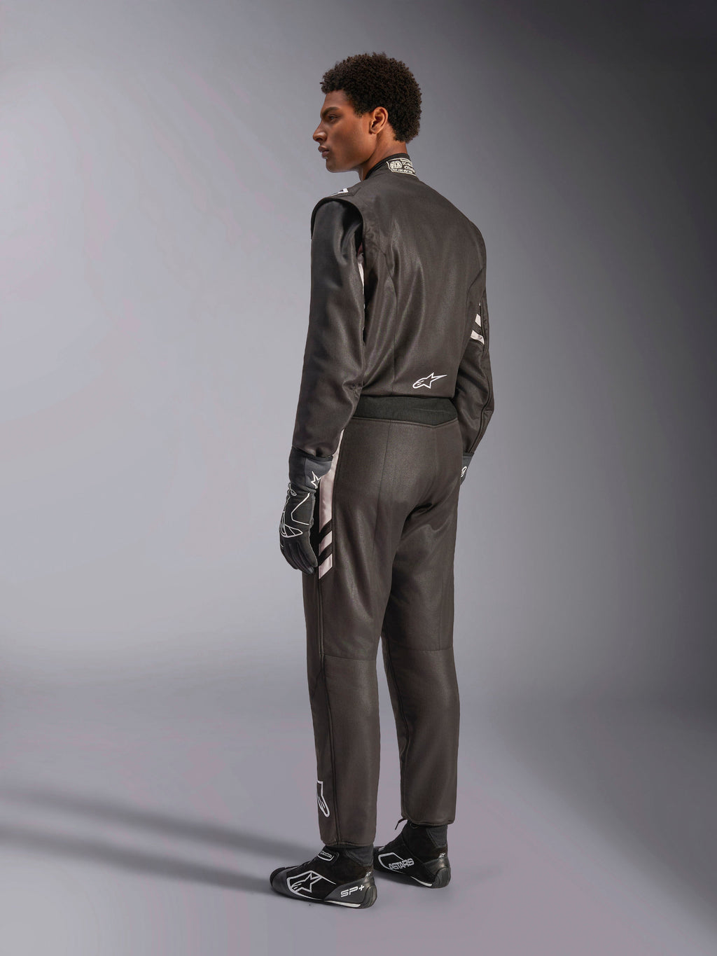 Alpinestars GP Race V3 Suit FIA, tuta da corsa automobilistica nera con dettagli argento, indossata da un modello in vista posteriore a tre quarti, abbinata a guanti e stivali da corsa neri coordinati, attrezzatura professionale da motorsport con design a due strati.