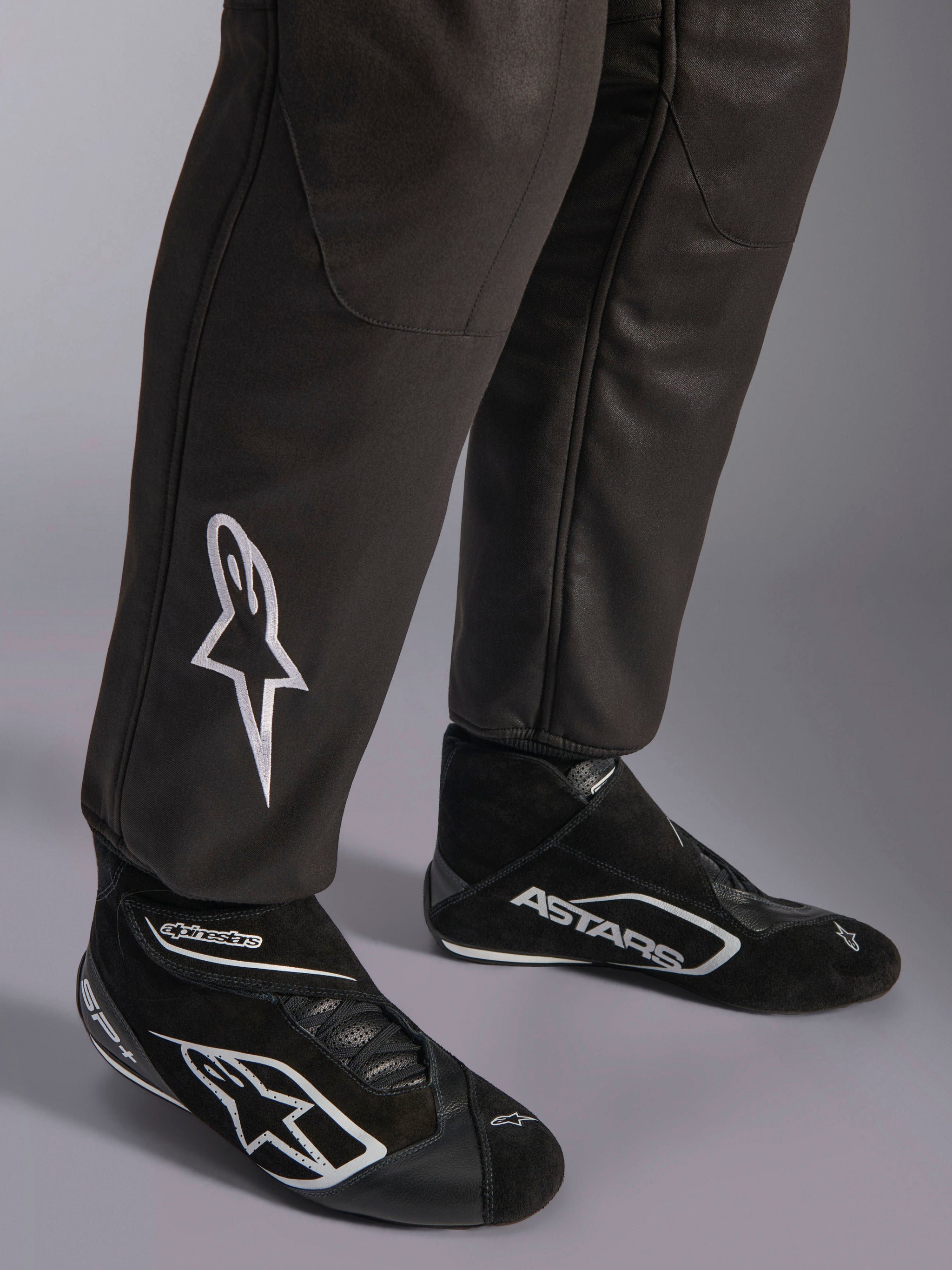 Alpinestars GP Race V3 Suit FIA, primo piano della parte inferiore delle gambe e degli stivali da corsa, nero con accenti argento, con logo a stella ricamato in bianco sullo stinco e polsini a coste alle caviglie, abbigliamento protettivo professionale per gare automobilistiche.