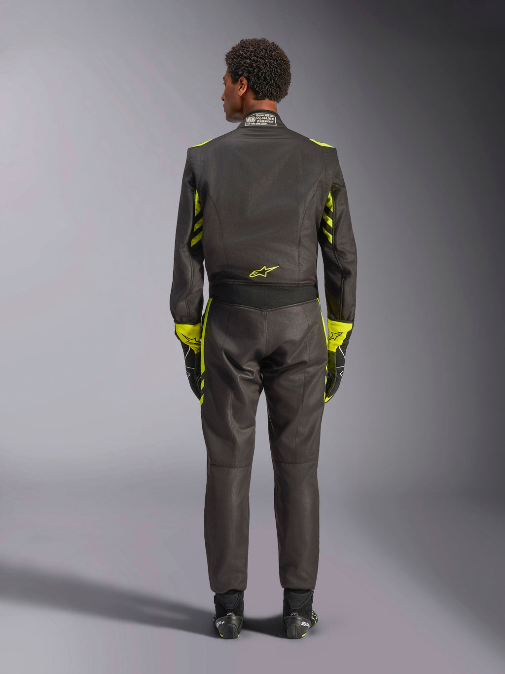 Alpinestars GP Race V3 Suit FIA, vista posteriore, nero con accenti giallo fluo, con pannelli gialli sulle braccia e sul busto, logo giallo sulla parte bassa della schiena, indossata con guanti e stivali da corsa abbinati, tuta professionale da corsa automobilistica
