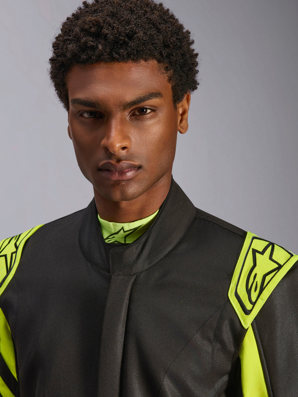 Primo piano di un modello che indossa la tuta Alpinestars GP Race V3 FIA in nero e giallo fluo, con spalline di estrazione gialle sulle spalle con loghi a stella, design a colletto alto, abbigliamento professionale per corse automobilistiche