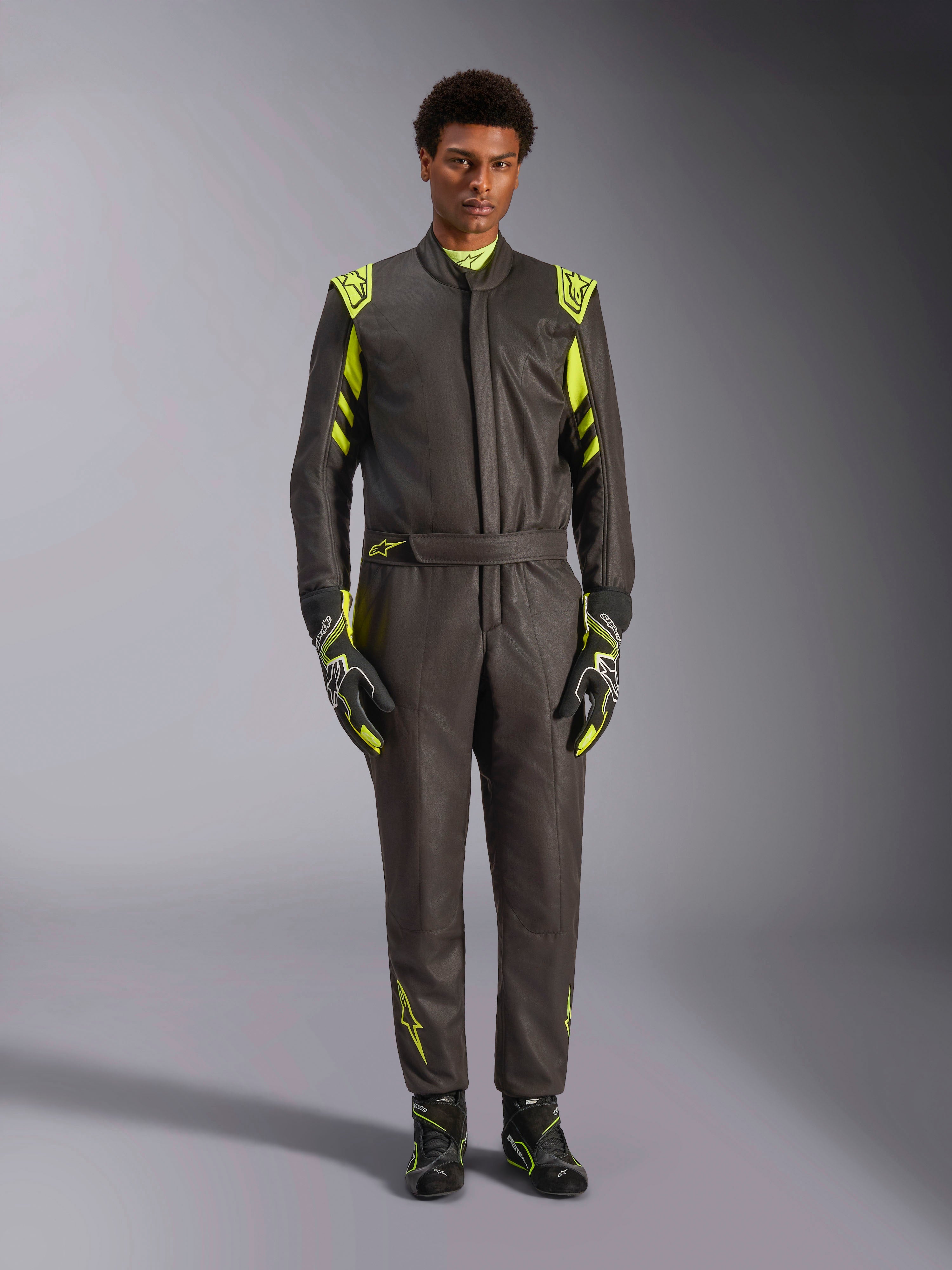 Tuta Alpinestars GP Race V3 FIA, tuta da auto racing, nero e giallo fluo, indossata da un modello con guanti e stivali da corsa abbinati, caratterizzata da accenti giallo fluo su spalle, vita e maniche, abbigliamento professionale per motorsport