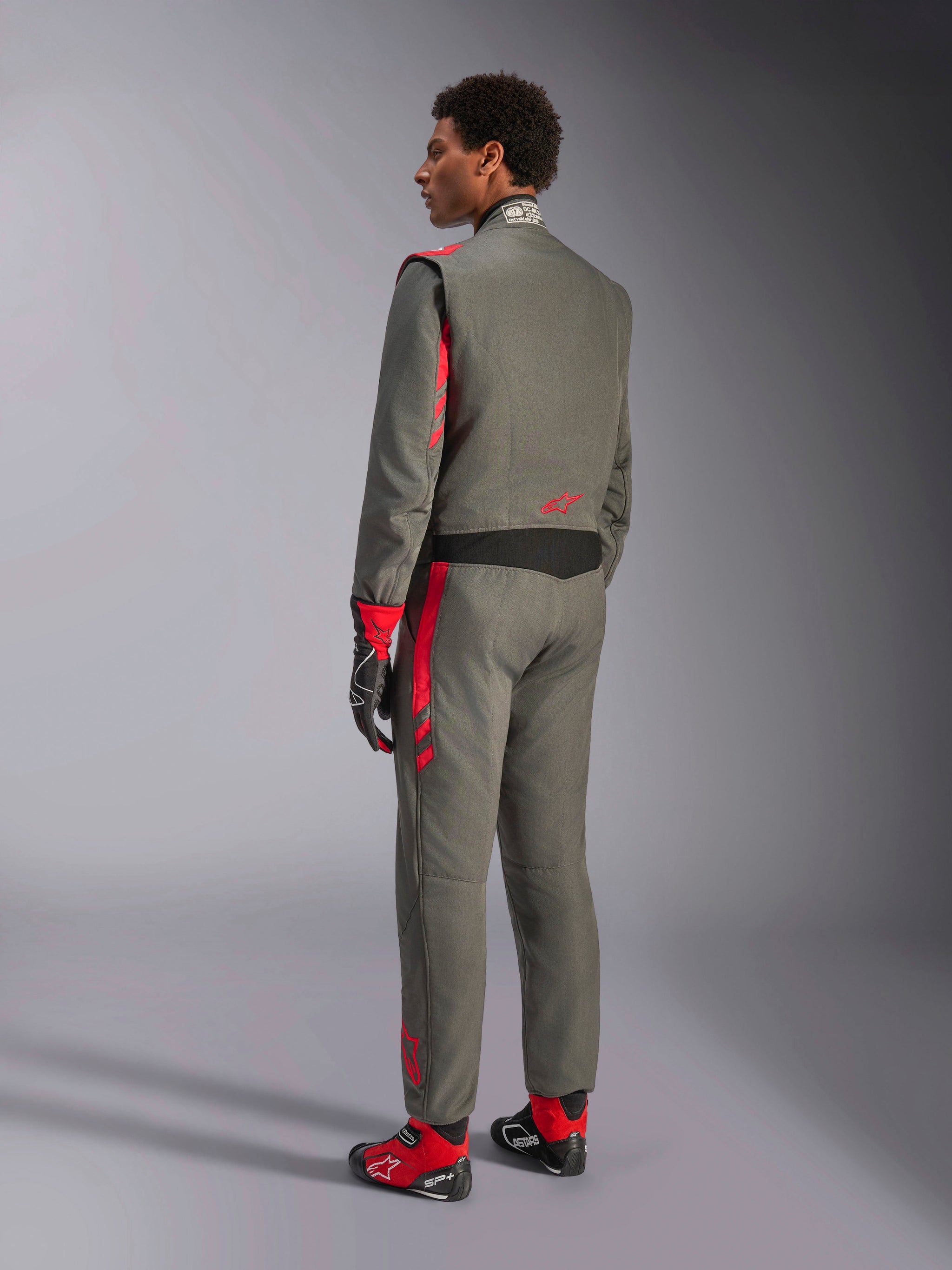 Alpinestars GP Race V3 Suit FIA, tuta da corsa grigio antracite e rosso, vista posteriore del modello che indossa la tuta a due strati, accenti rossi su spalle e gambe, abbinata a guanti e stivali coordinati, equipaggiamento professionale per gare automobilistiche