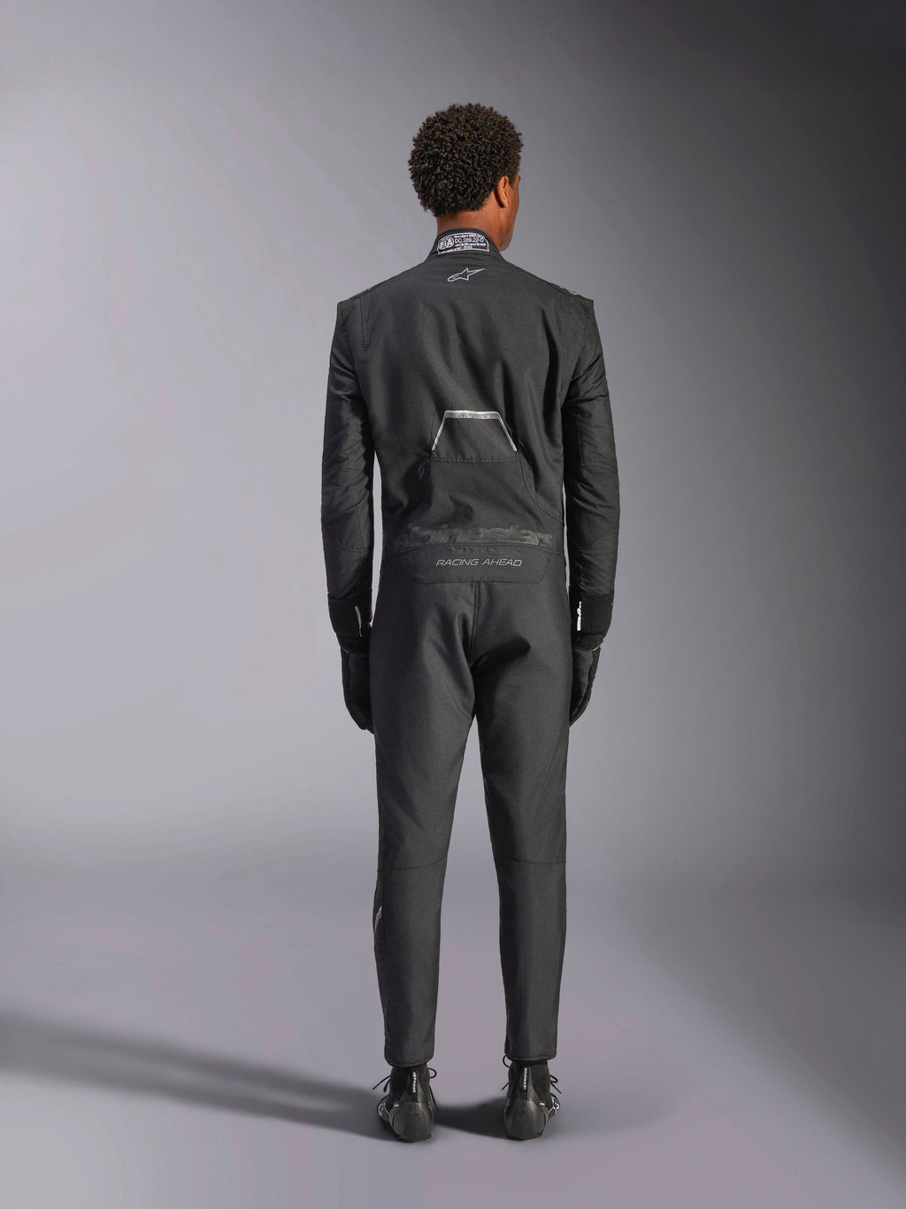 Tuta Alpinestars Hypertech V3, Black Dark Shadow, vista posteriore di una tuta intera professionale da auto racing, con etichetta di certificazione FIA sul colletto e marchi 'Alpinestars' e 'RACING AHEAD' sulla zona lombare, indossata con guanti e stivali da corsa neri coordinati.