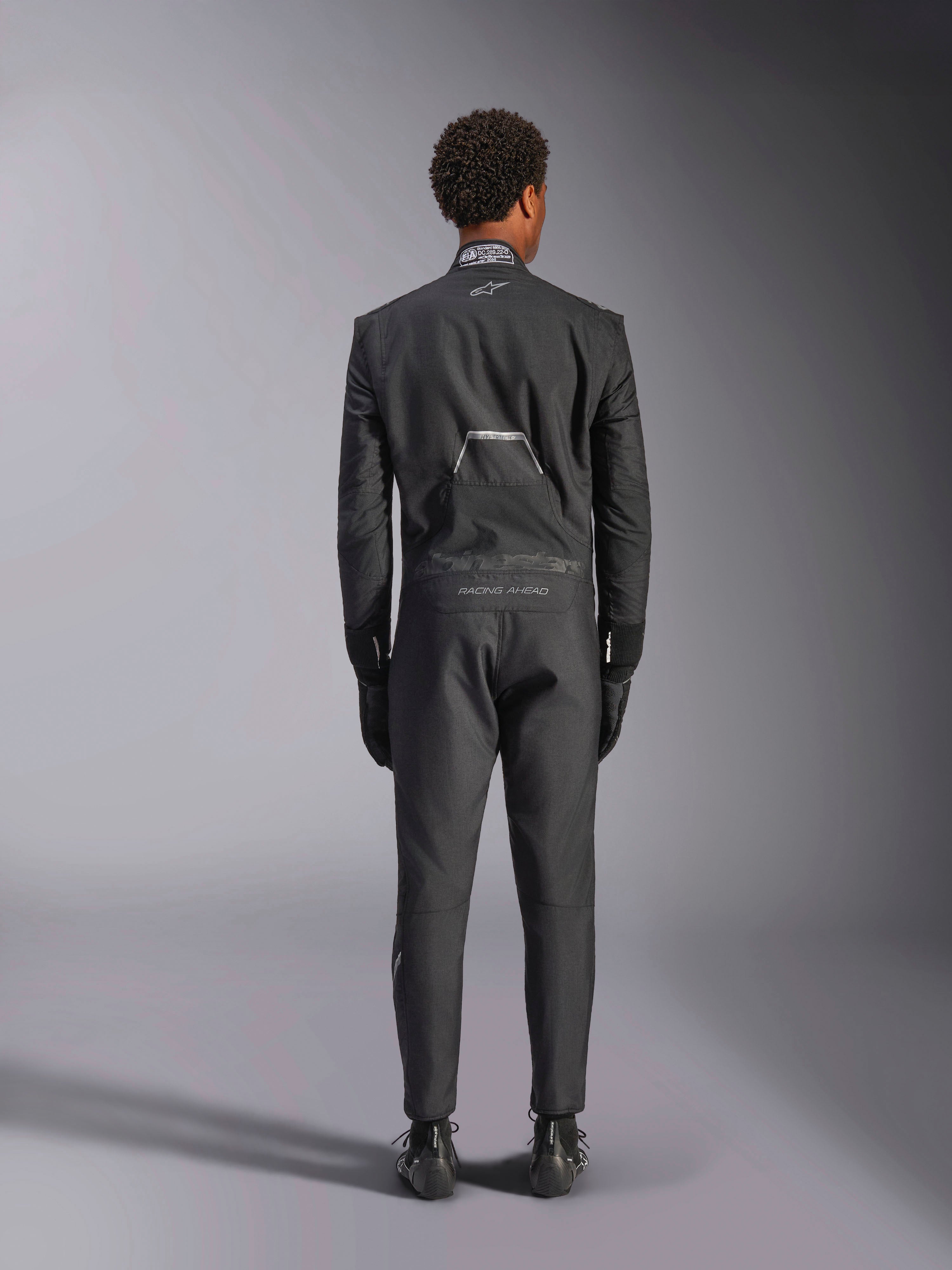 Tuta Alpinestars Hypertech V3, Black Dark Shadow, vista posteriore di una tuta intera professionale da auto racing, con etichetta di certificazione FIA sul colletto e marchi 'Alpinestars' e 'RACING AHEAD' sulla zona lombare, indossata con guanti e stivali da corsa neri coordinati.