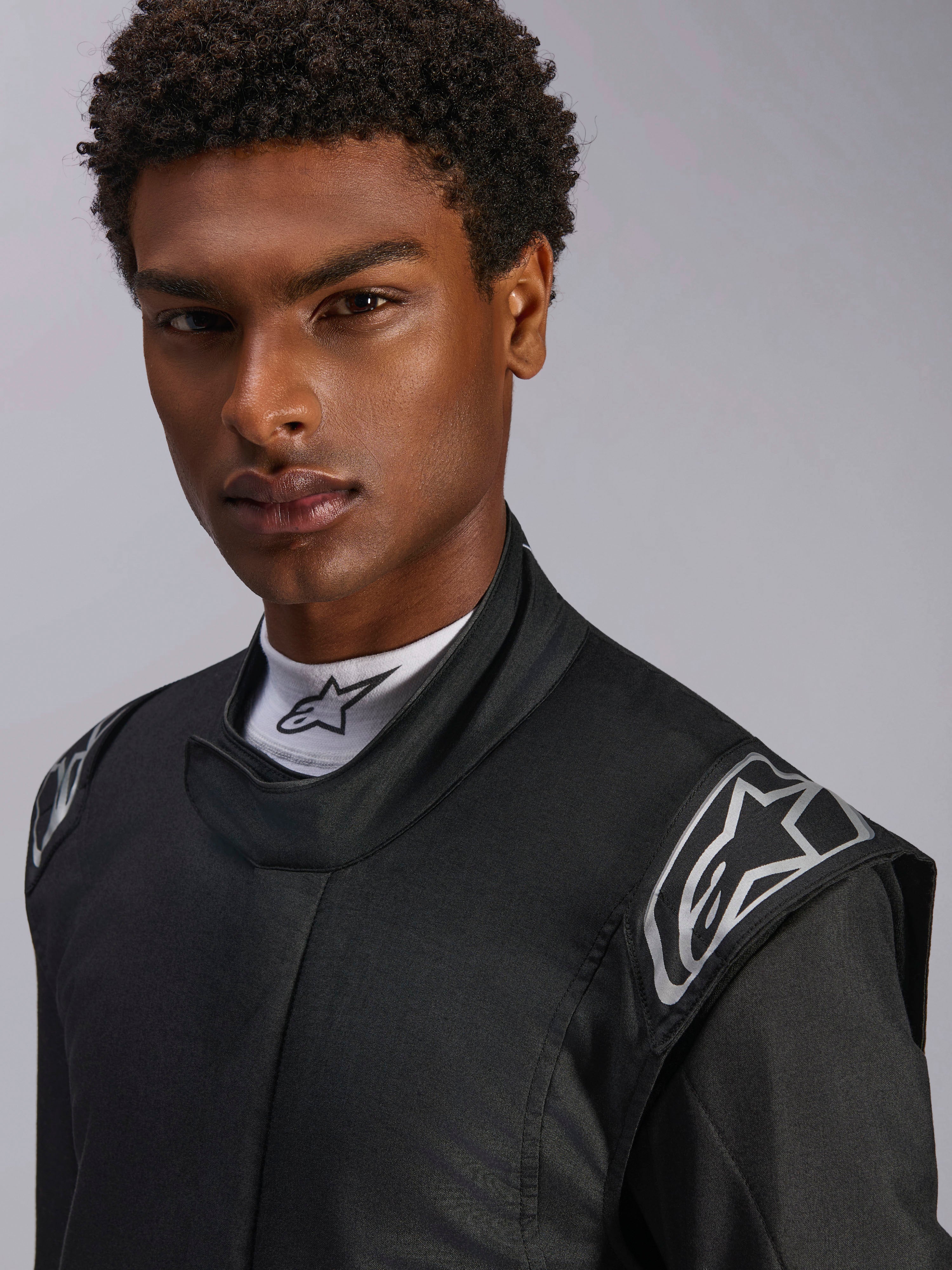 Tuta Alpinestars Hypertech V3, tuta da auto racing nero Dark Shadow, primo piano su modello, loghi argentati sulle spalle, colletto alto con maglia a lupetto bianca Alpinestars, abbigliamento protettivo professionale per motorsport