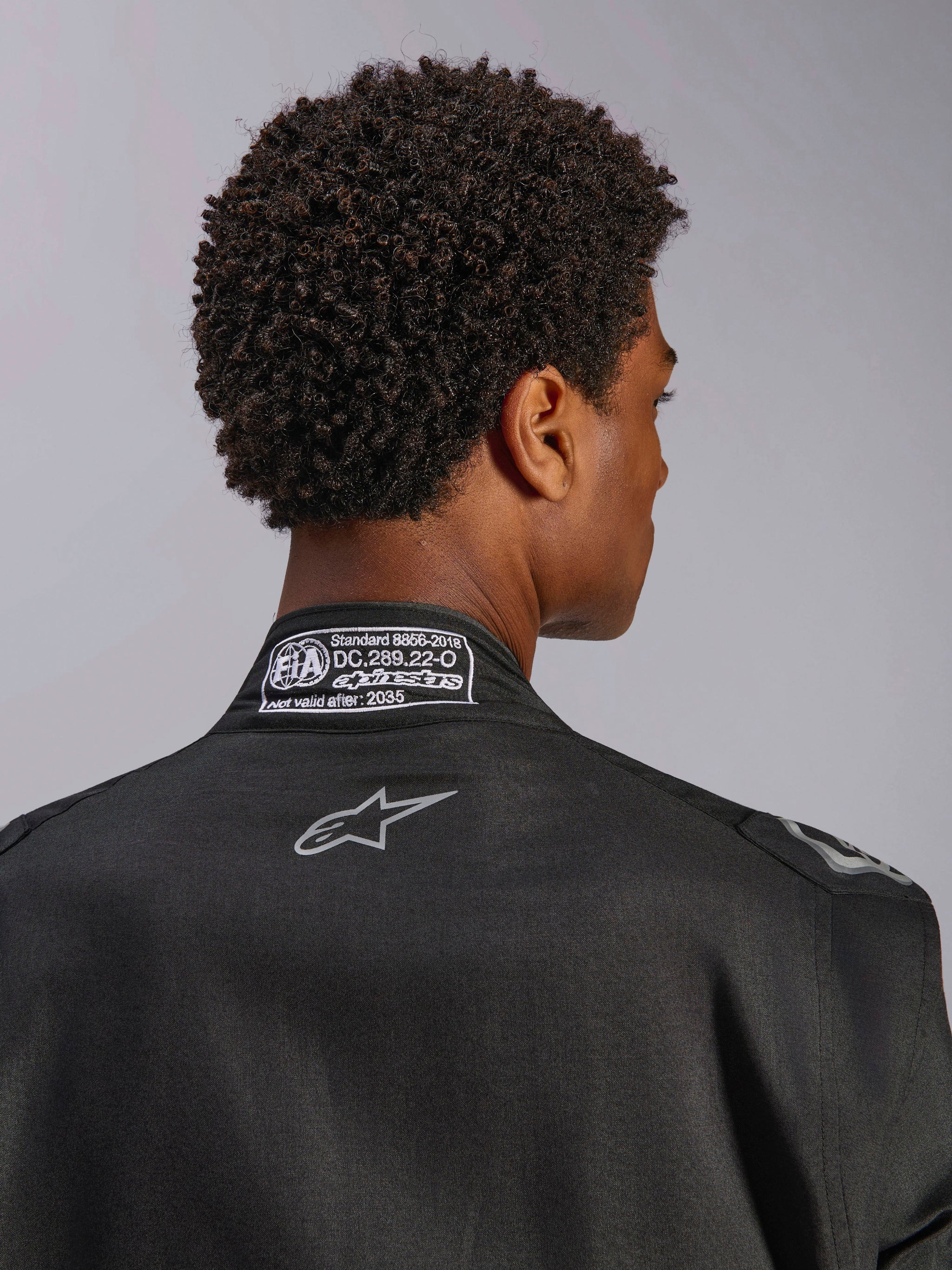 Tuta Alpinestars Hypertech V3, Black Dark Shadow, vista posteriore ravvicinata del colletto con etichetta di certificazione FIA 8856-2018 e logo sulla schiena, tuta da corsa professionale leggera con vestibilità atletica