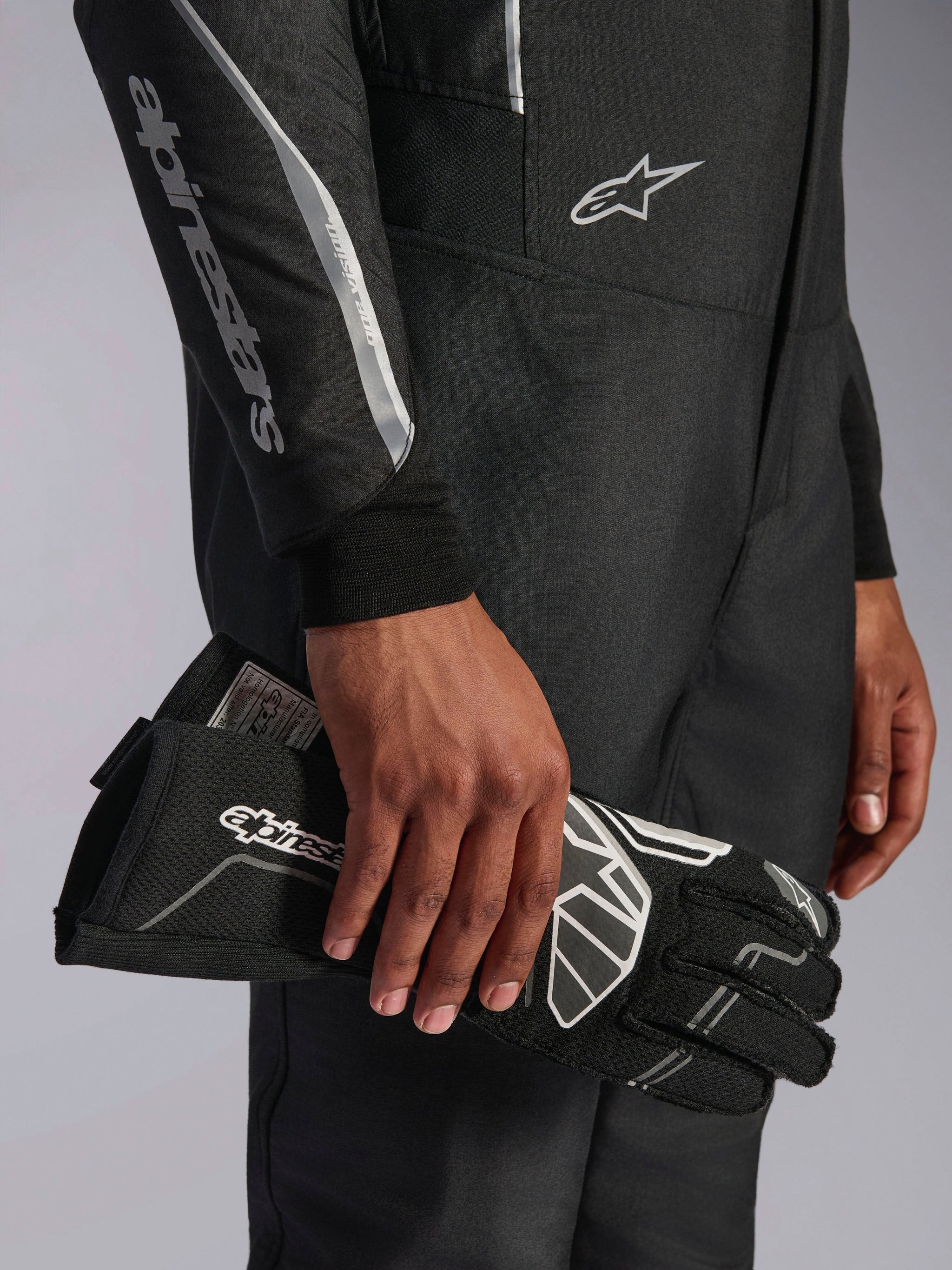 Tuta Alpinestars Hypertech V3 in Black Dark Shadow, dettaglio ravvicinato della manica e del busto con logo Alpinestars argento, mano che tiene un guanto da corsa nero con accenti bianchi, equipaggiamento professionale da auto racing
