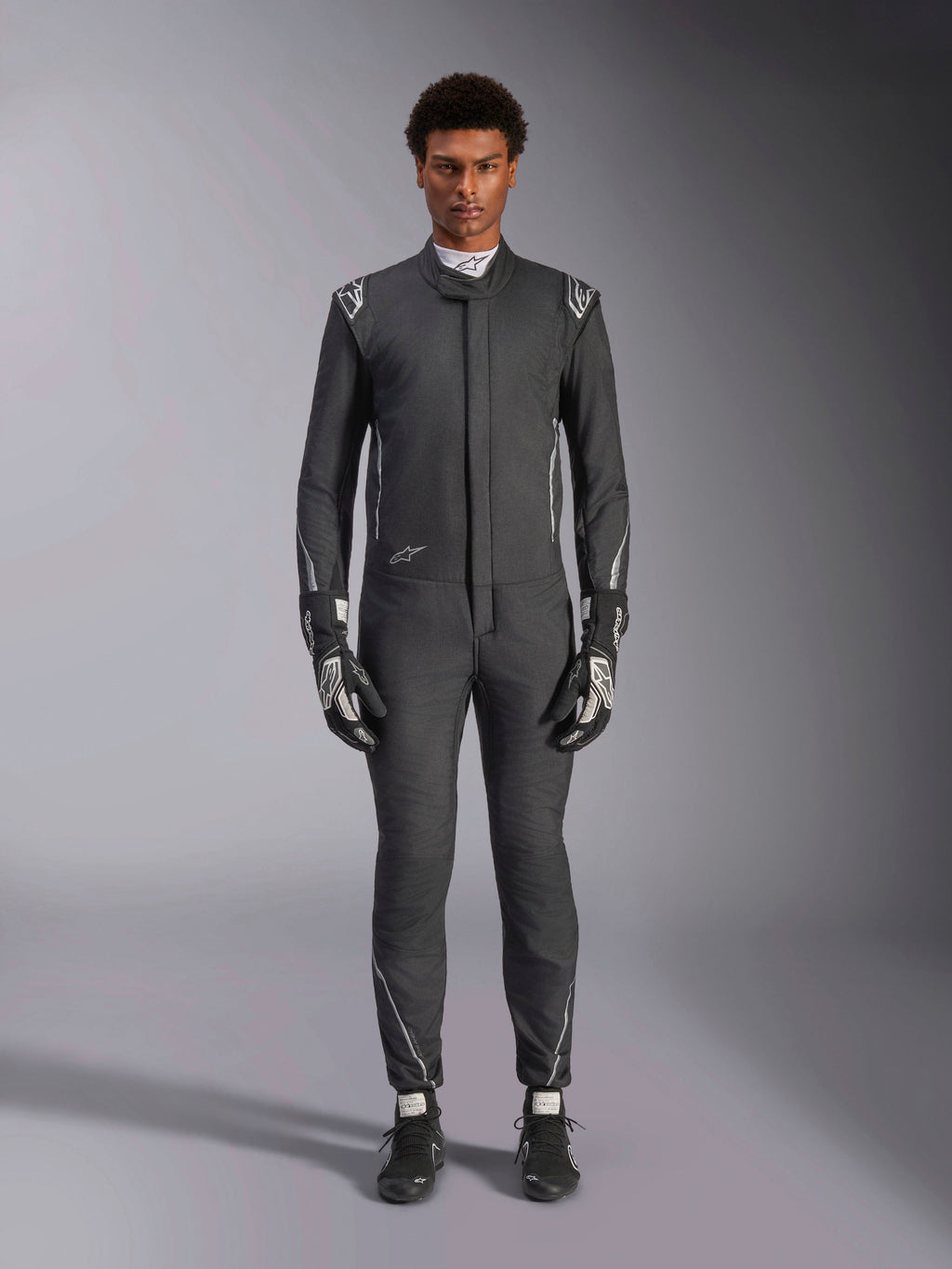 Tuta Alpinestars Hypertech V3, tuta da corsa professionale, nero e Dark Shadow, indossata da un modello con guanti e scarpe da corsa coordinati, vestibilità atletica aderente con accenti argento e loghi, abbigliamento motorsport ad alte prestazioni