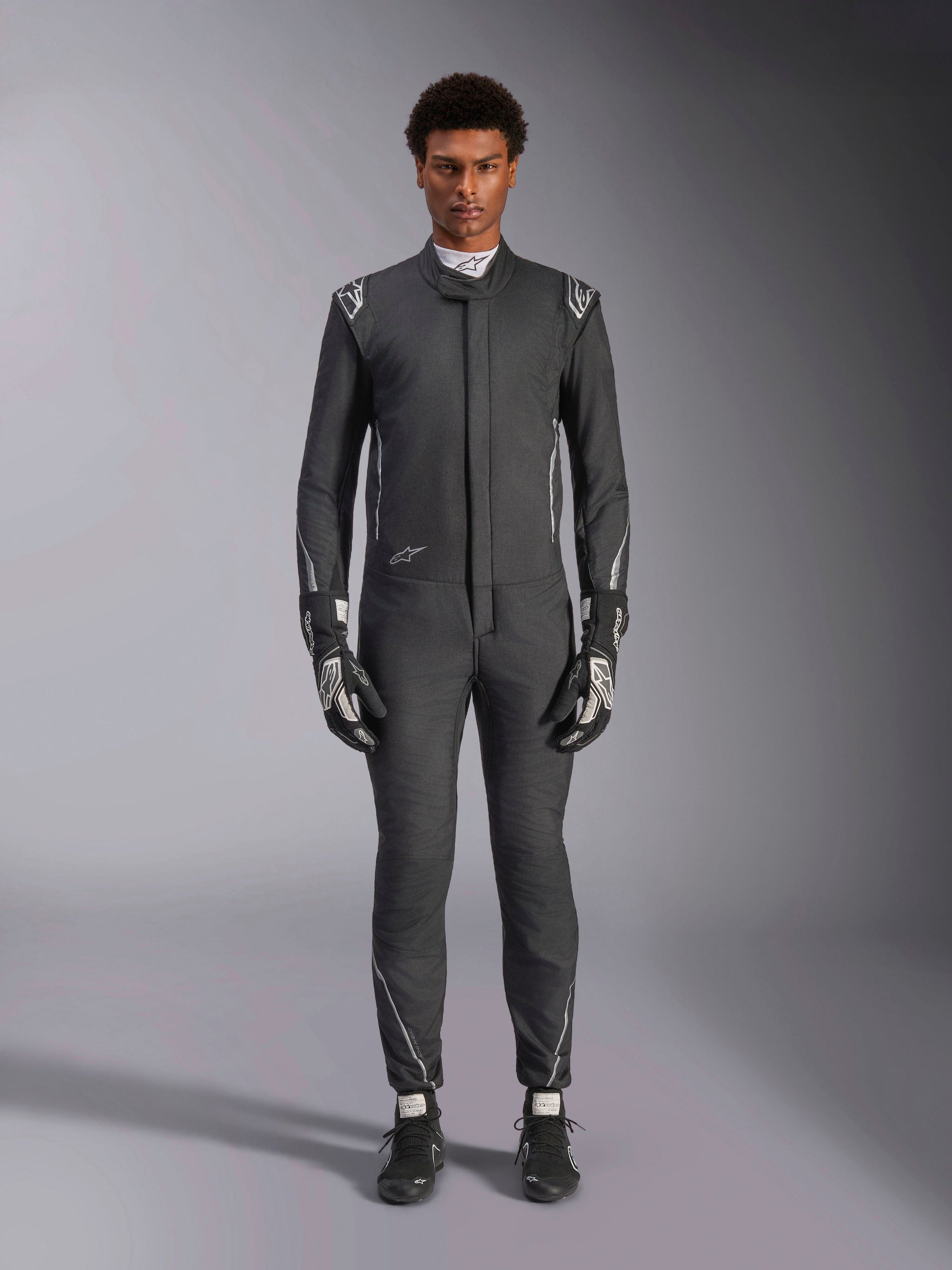 Tuta Alpinestars Hypertech V3, tuta da corsa professionale, nero e Dark Shadow, indossata da un modello con guanti e scarpe da corsa coordinati, vestibilità atletica aderente con accenti argento e loghi, abbigliamento motorsport ad alte prestazioni