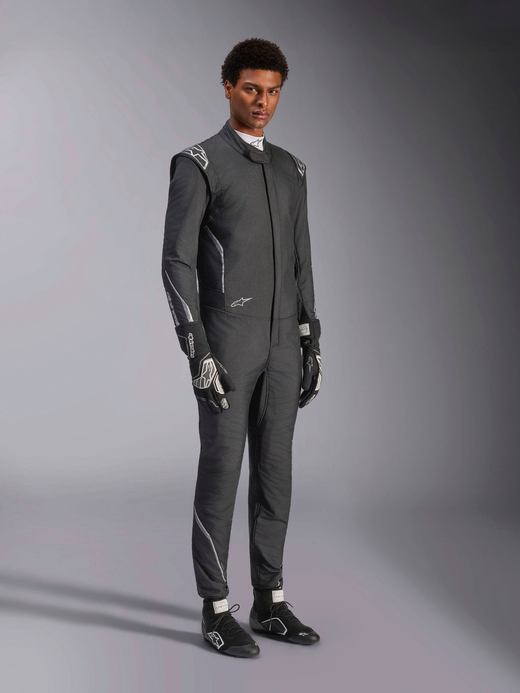 Tuta Alpinestars Hypertech V3, tuta da auto racing, nero e Dark Shadow, indossata da un modello con guanti neri e scarpe da corsa coordinati, vestibilità slim atletica, loghi Alpinestars e accenti grigio chiaro, abbigliamento protettivo professionale per motorsport