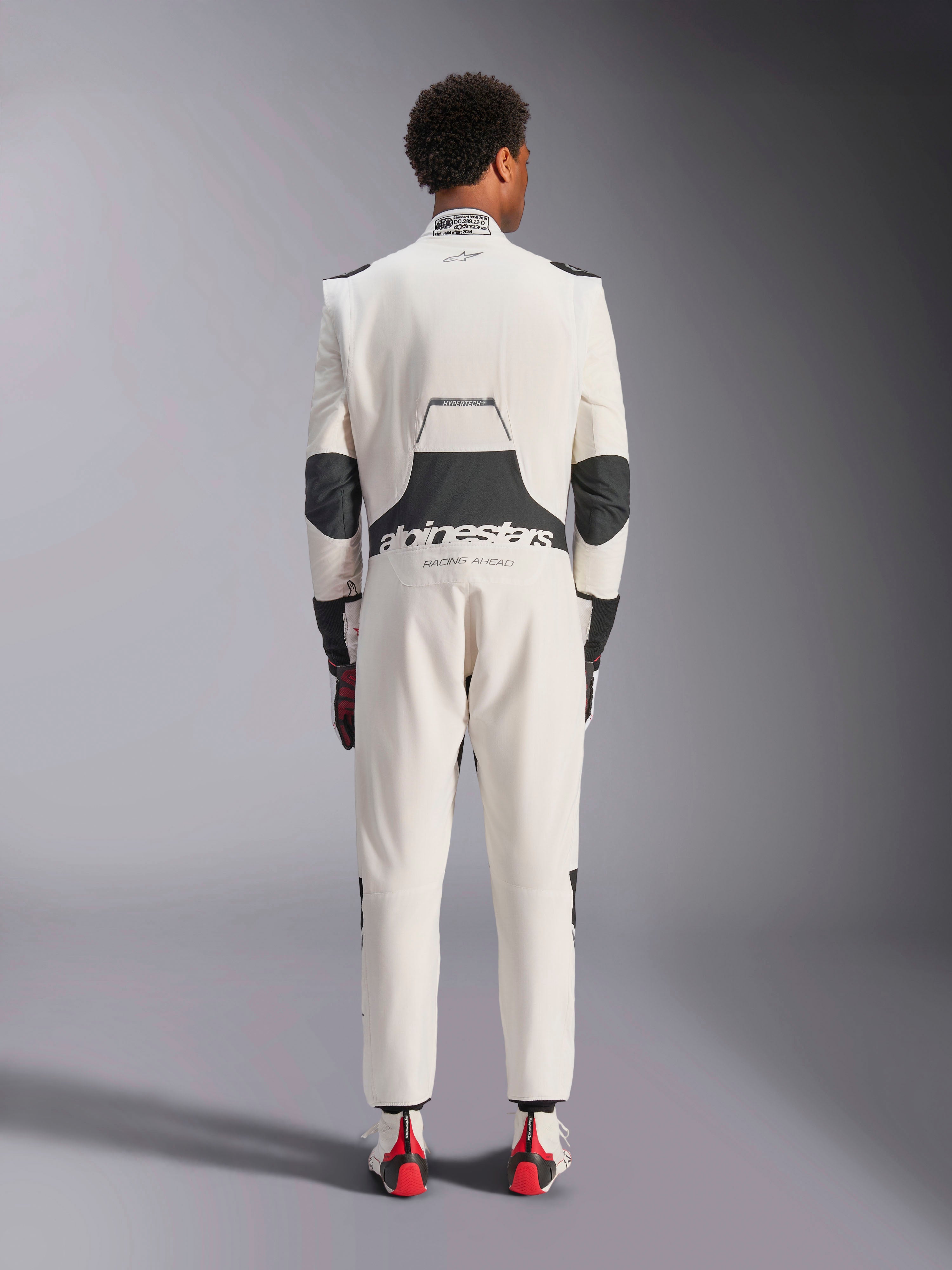 Vista posteriore di un modello che indossa la tuta Alpinestars Hypertech V3 in White Black Silver, che mette in risalto l'ampio pannello elasticizzato lombare nero con i loghi 'alpinestars' e 'RACING AHEAD', inserti neri sui gomiti, l'etichetta di certificazione FIA sul colletto e una vestibilità atletica e aderente progettata per le competizioni professionali.