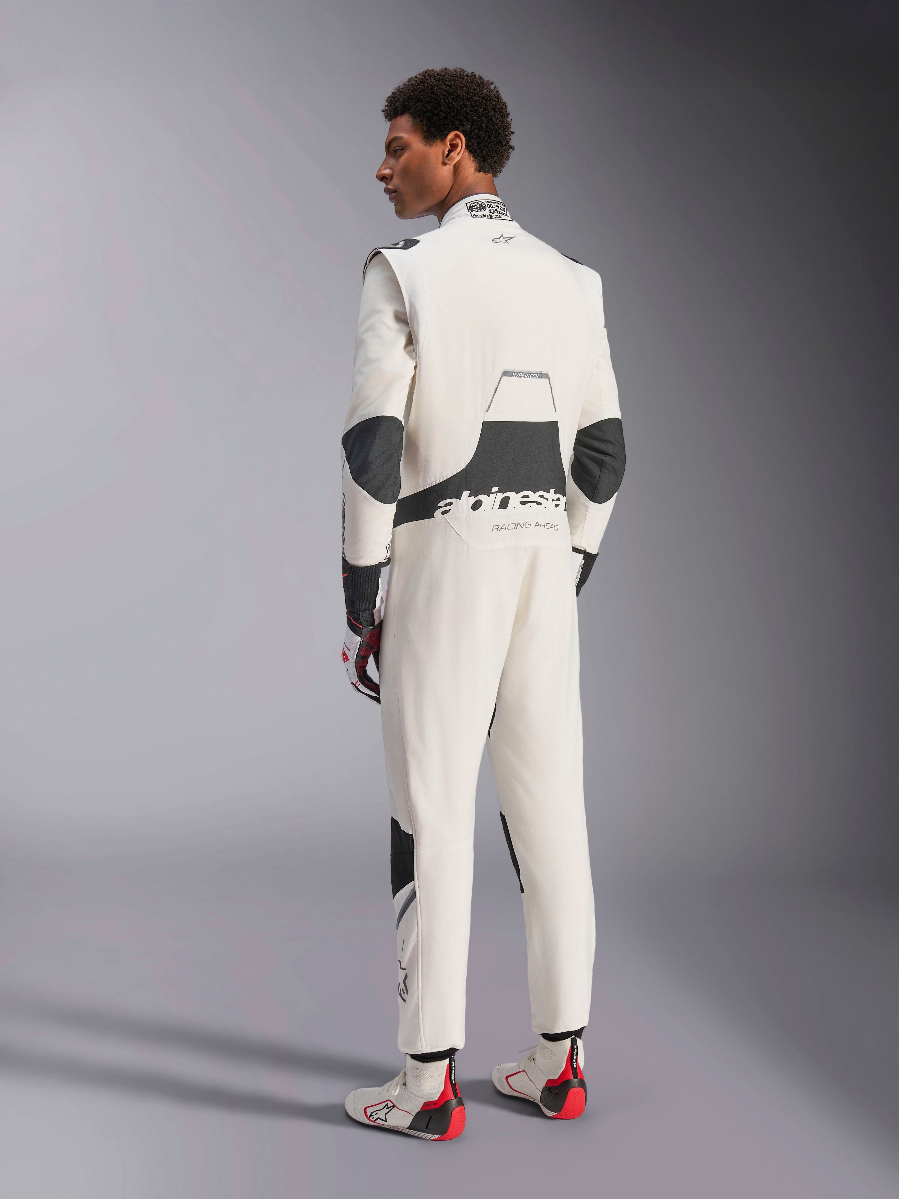 Tuta Alpinestars Hypertech V3, tuta da corsa professionale, bianco, nero e argento, vista posteriore del modello che mostra i pannelli elasticizzati neri sulla zona lombare e sui gomiti, marchio Alpinestars sulla schiena, abbinata a guanti e stivali da gara coordinati per un look da motorsport completo