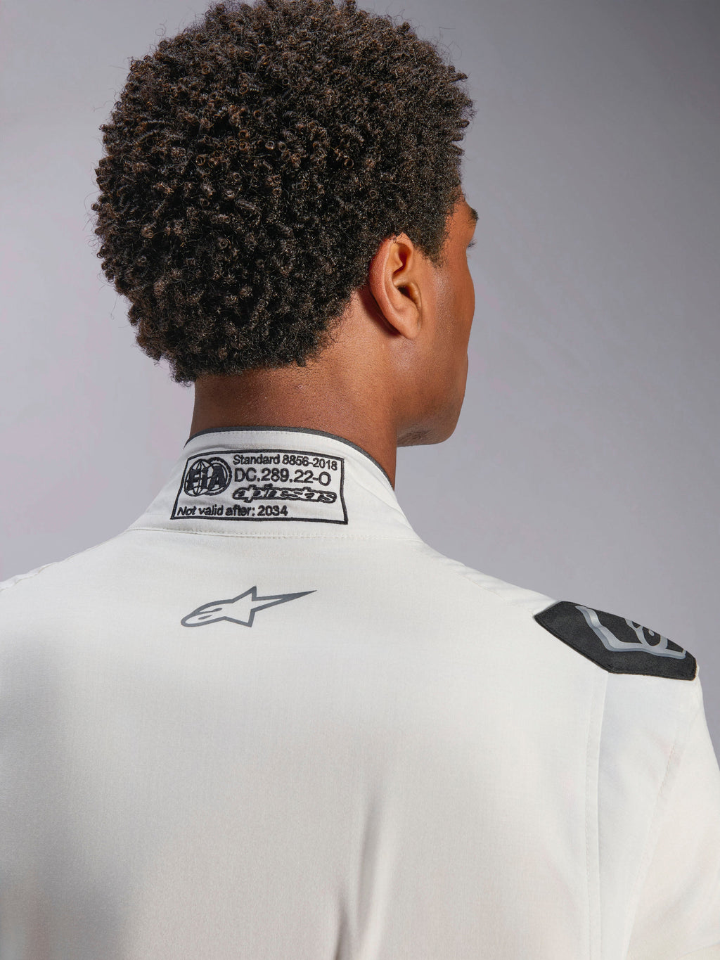 Tuta Alpinestars Hypertech V3, bianca, vista dettagliata del colletto posteriore e della spalla, con etichetta di certificazione FIA 8856-2018, logo Alpinestars grigio stampato e maniglia di estrazione nera, abbigliamento da corsa professionale