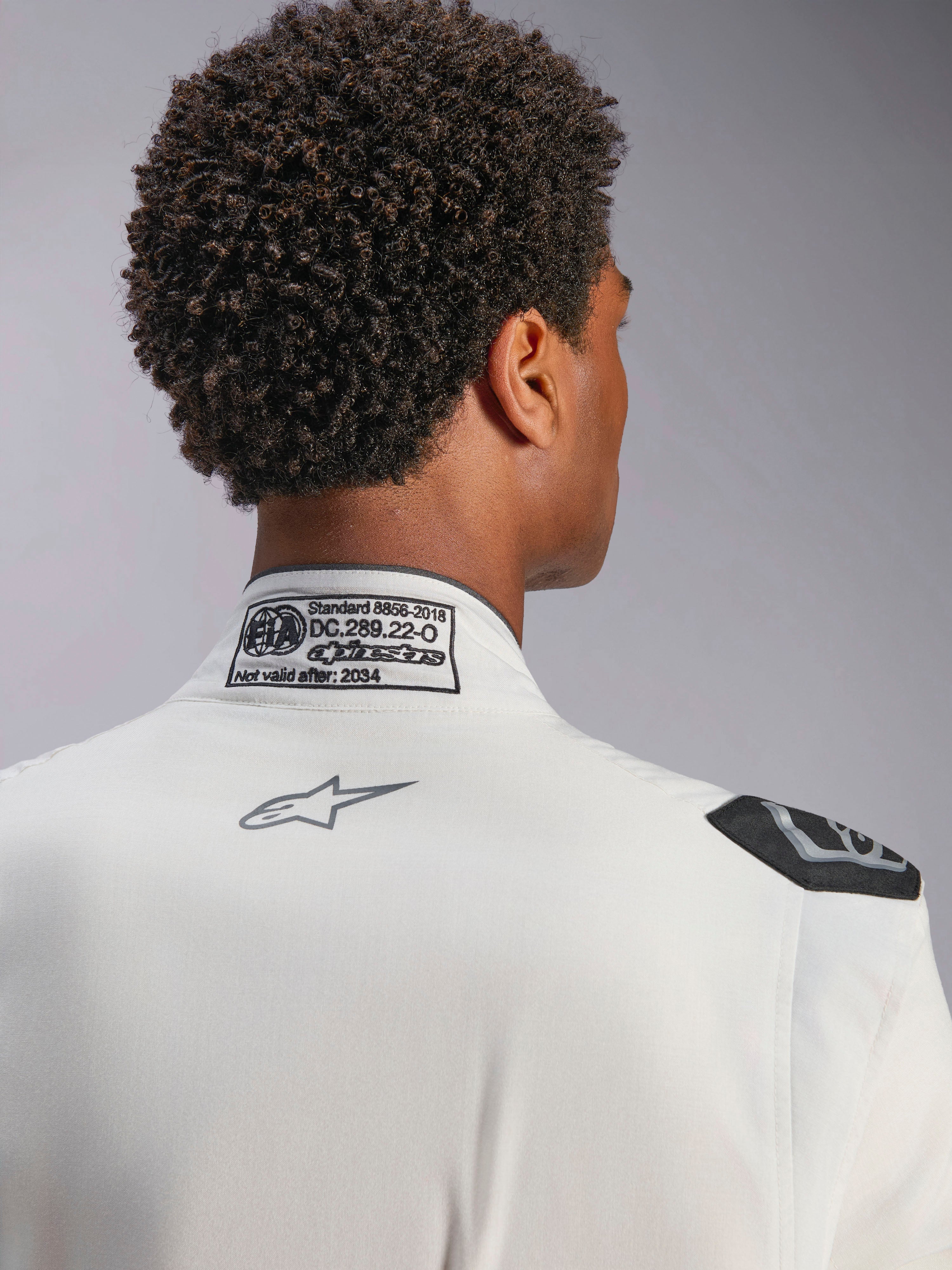 Tuta Alpinestars Hypertech V3, bianca, vista dettagliata del colletto posteriore e della spalla, con etichetta di certificazione FIA 8856-2018, logo Alpinestars grigio stampato e maniglia di estrazione nera, abbigliamento da corsa professionale