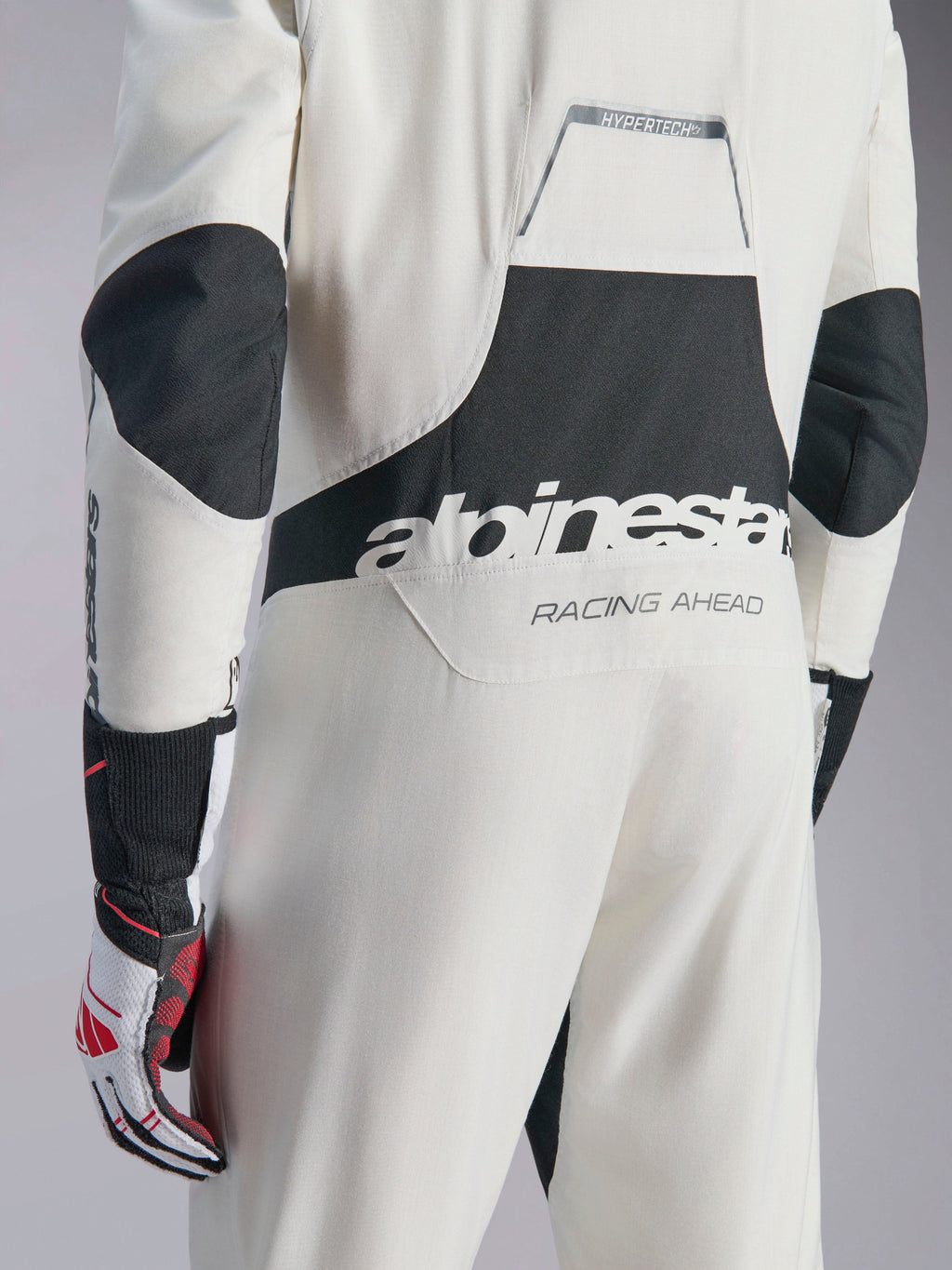 Tuta Alpinestars Hypertech V3, tuta da auto racing bianca, nera e argento, vista posteriore con grande logo 'alpinestars' nero e testo 'RACING AHEAD', con pannelli elasticizzati neri su gomiti e zona lombare, abbigliamento protettivo professionale da motorsport.