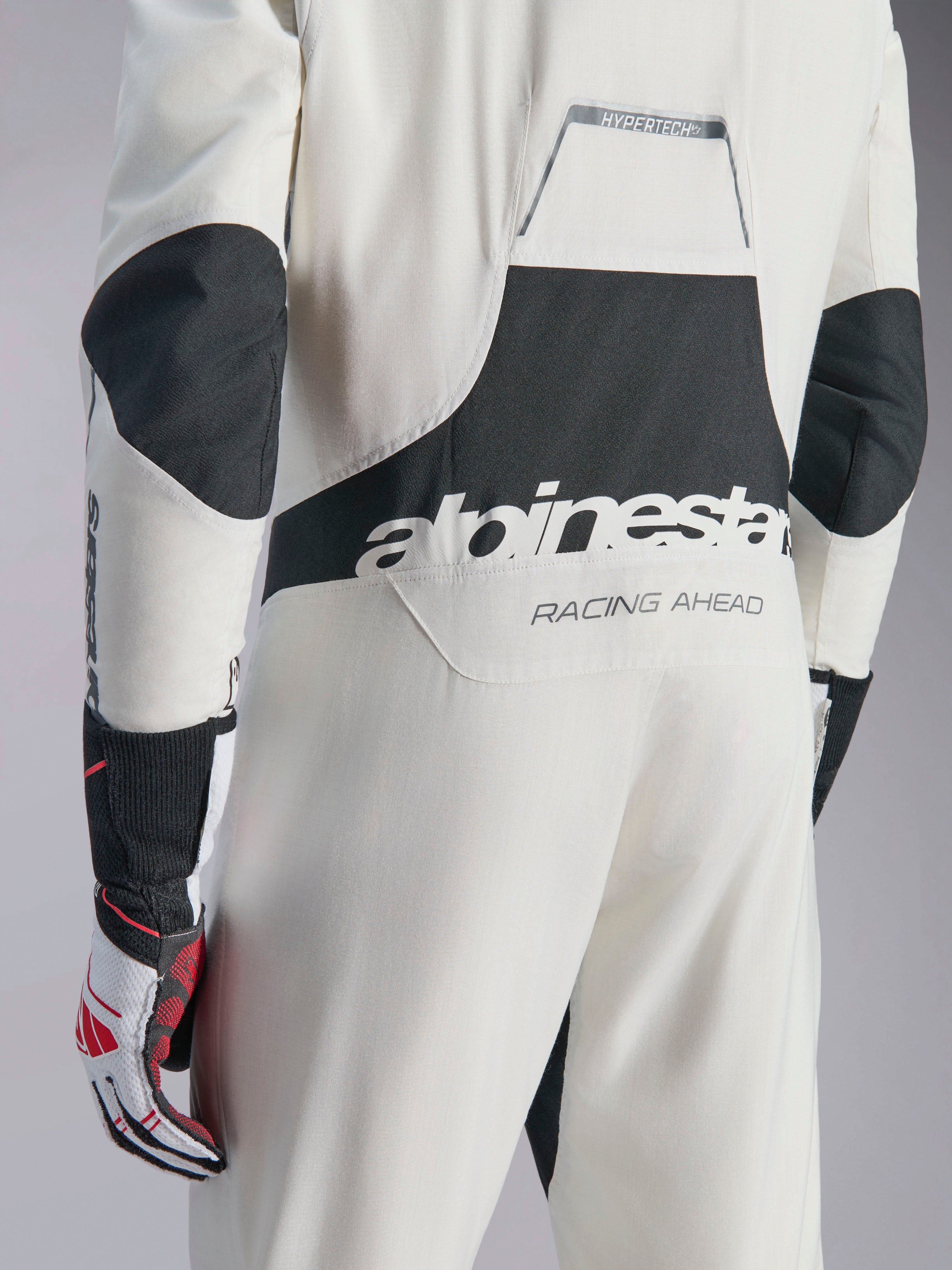 Tuta Alpinestars Hypertech V3, tuta da auto racing bianca, nera e argento, vista posteriore con grande logo 'alpinestars' nero e testo 'RACING AHEAD', con pannelli elasticizzati neri su gomiti e zona lombare, abbigliamento protettivo professionale da motorsport.