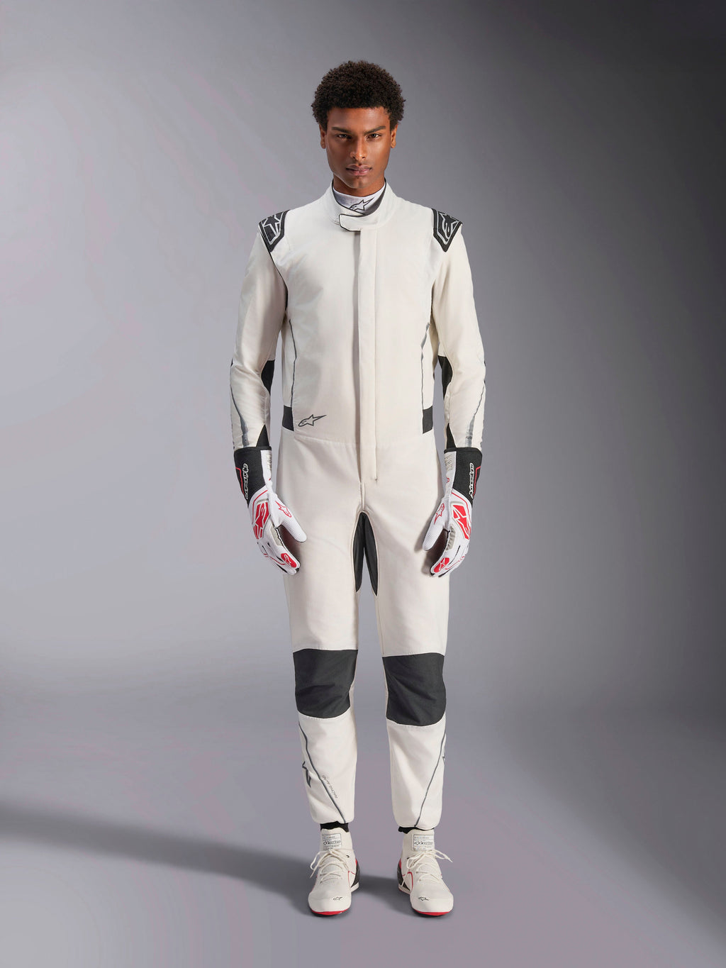 Tuta Alpinestars Hypertech V3, tuta professionale da auto racing, bianco, nero e argento, indossata da un modello con guanti rossi e bianchi e scarpe da racing bianche coordinate, caratterizzata da pannelli neri a contrasto e loghi Alpinestars, design con vestibilità atletica slim fit