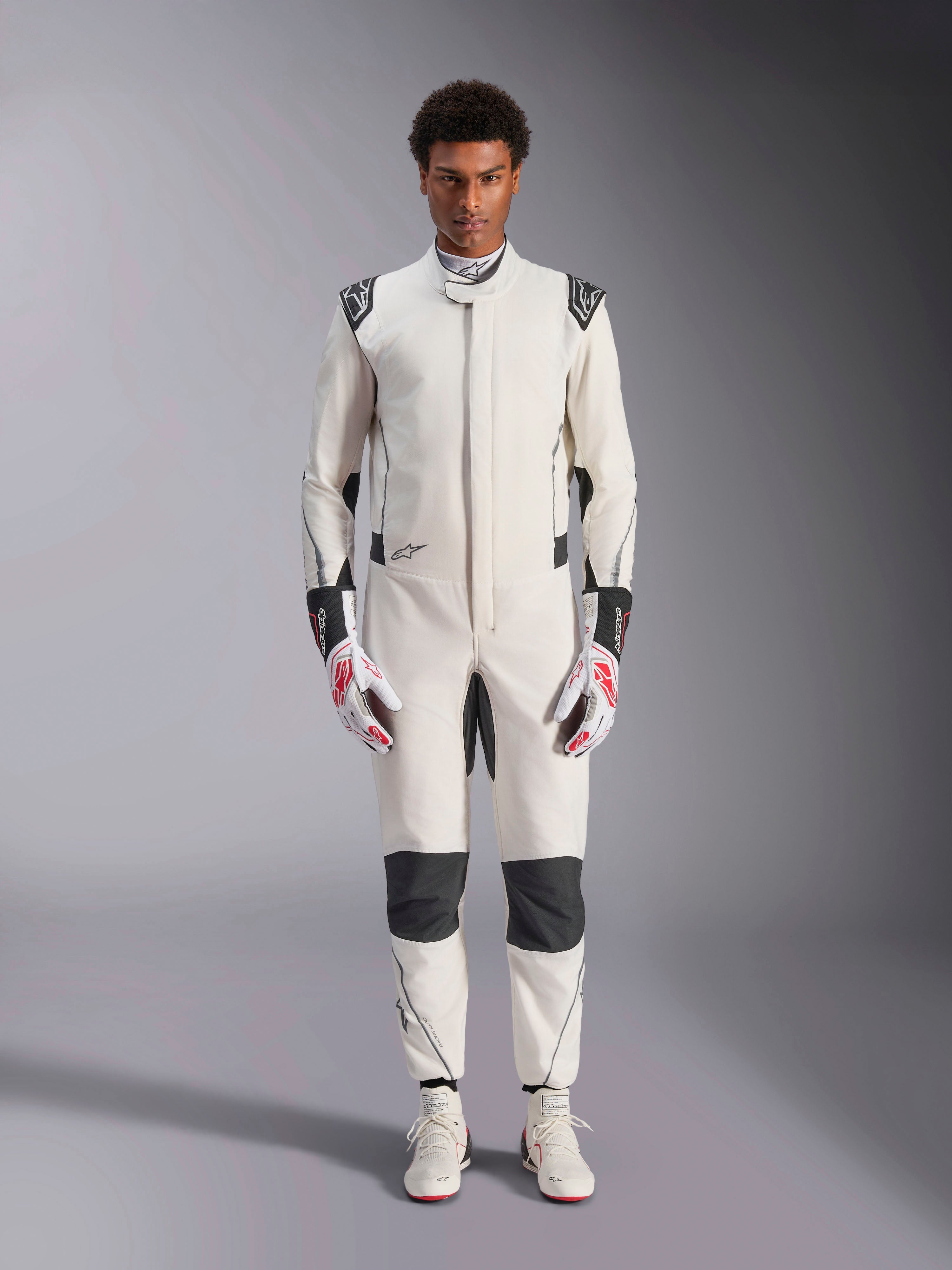 Tuta Alpinestars Hypertech V3, tuta professionale da auto racing, bianco, nero e argento, indossata da un modello con guanti rossi e bianchi e scarpe da racing bianche coordinate, caratterizzata da pannelli neri a contrasto e loghi Alpinestars, design con vestibilità atletica slim fit