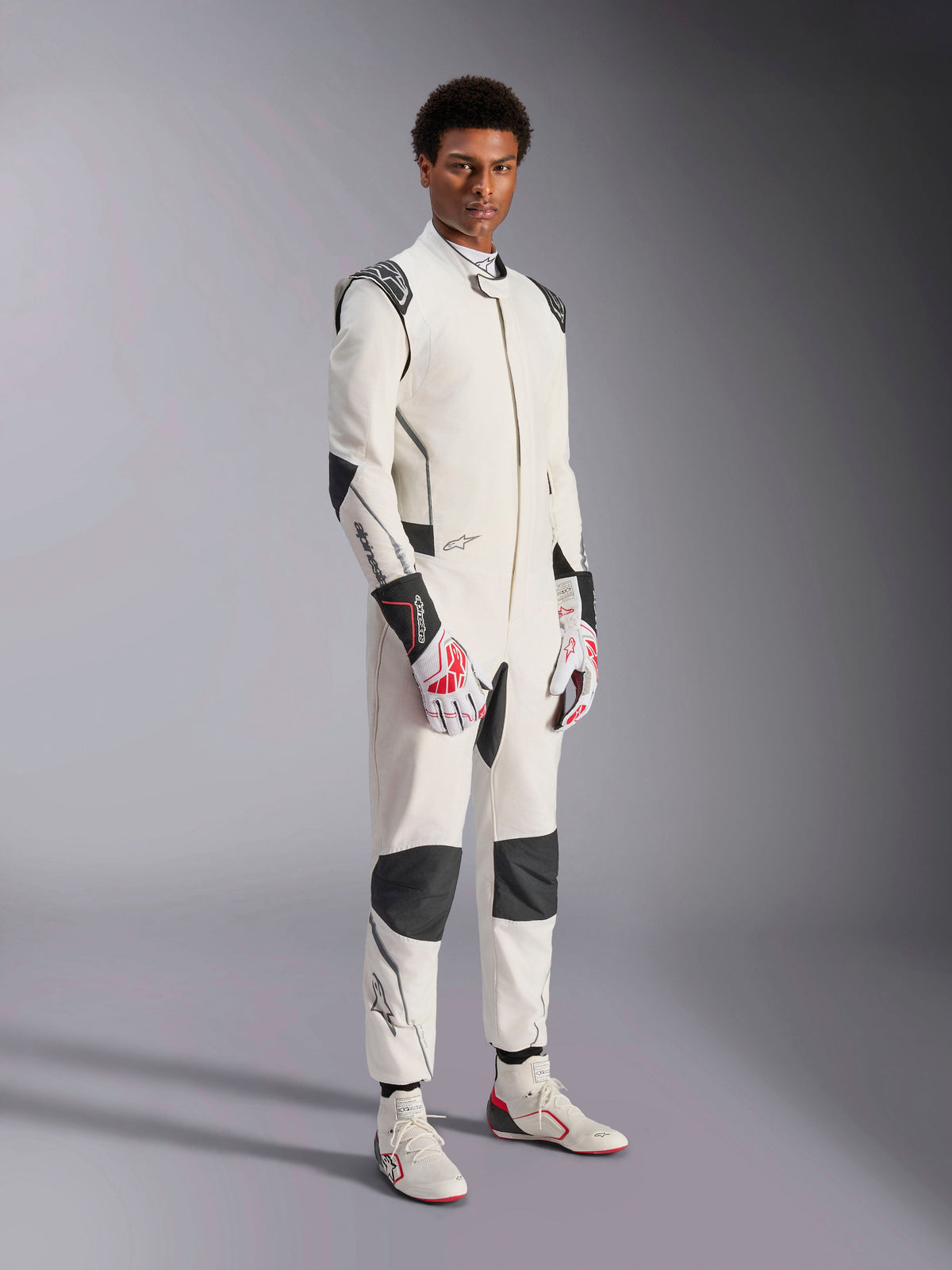 Modello che indossa la tuta Alpinestars Hypertech V3, bianco con accenti neri e argento, abbigliamento professionale per corse automobilistiche con pannelli neri su spalle e ginocchia, abbinata a guanti da corsa bianchi e rossi e stivali da corsa bianchi