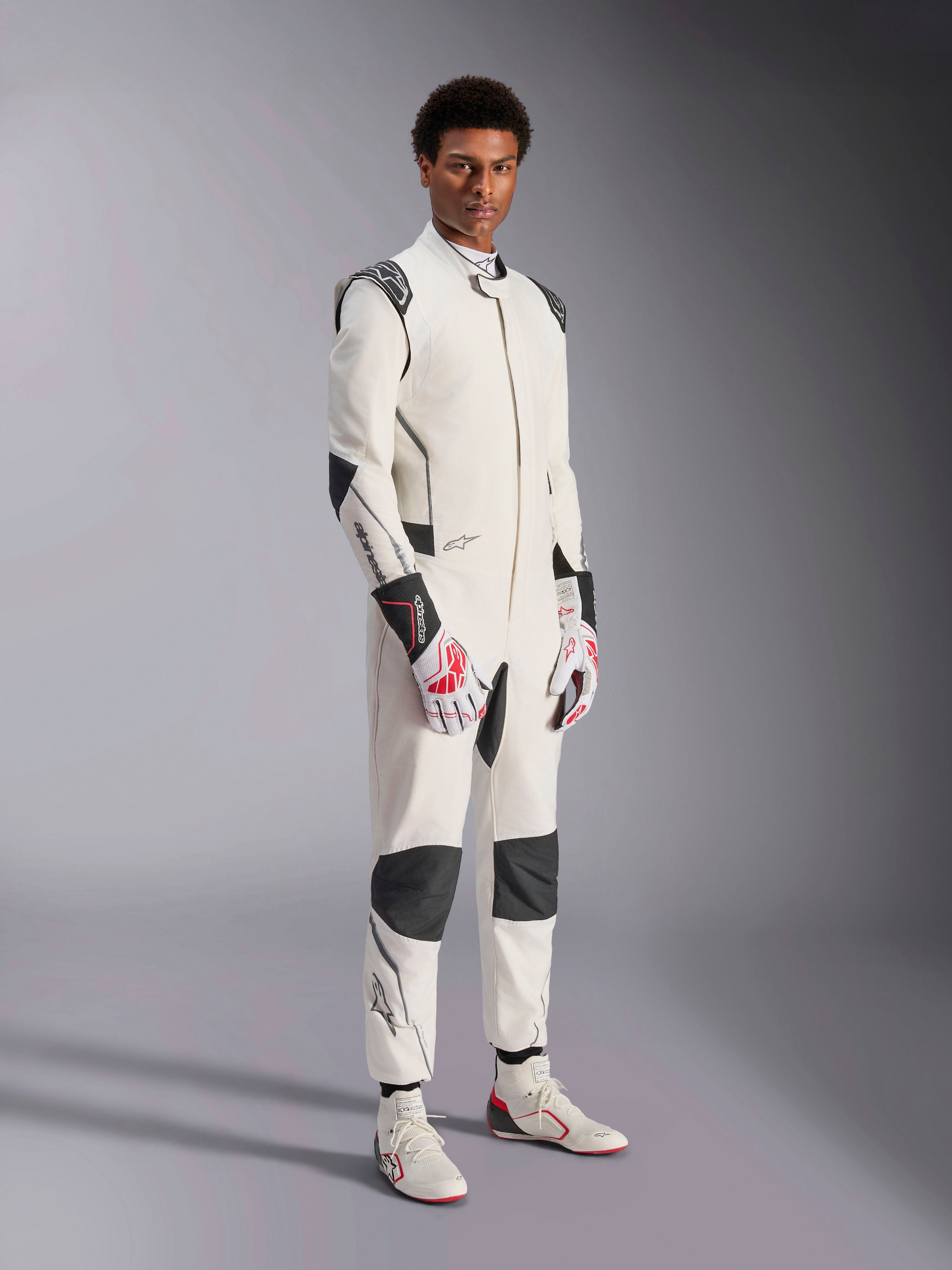 Modello che indossa la tuta Alpinestars Hypertech V3, bianco con accenti neri e argento, abbigliamento professionale per corse automobilistiche con pannelli neri su spalle e ginocchia, abbinata a guanti da corsa bianchi e rossi e stivali da corsa bianchi