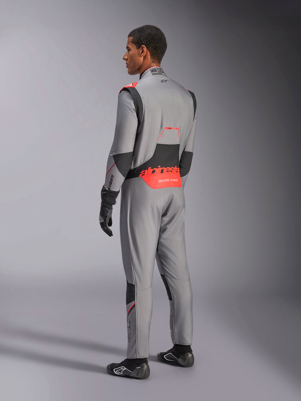 Tuta Alpinestars Hypertech V3 in grigio medio, nero e rosso fluo, vista posteriore di tre quarti, con logo Alpinestars rosso sulla parte bassa della schiena, pannelli elasticizzati neri e vestibilità atletica per competizioni automobilistiche professionali.