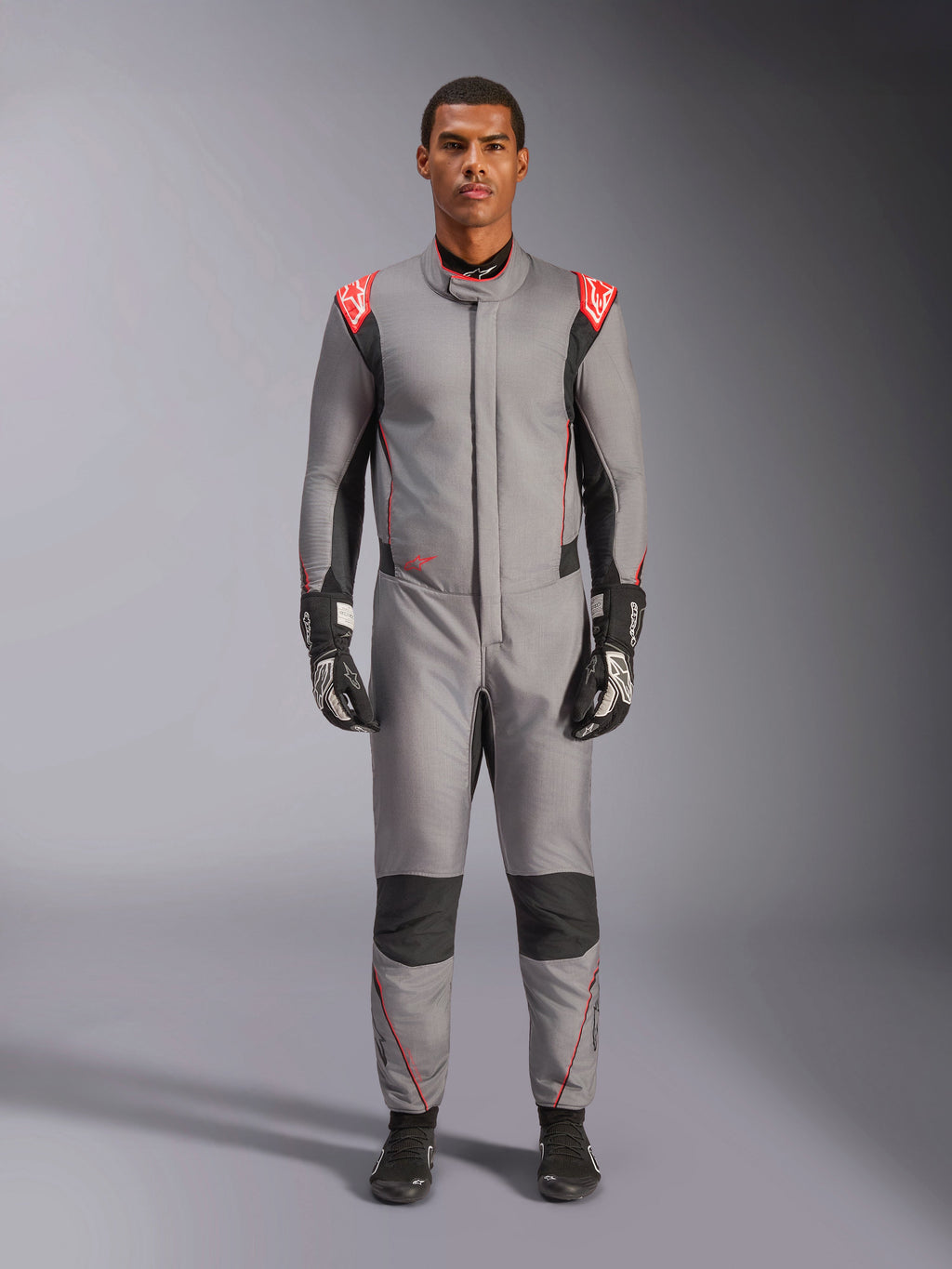 Alpinestars Hypertech V3 Suit, tuta da corsa professionale in grigio medio, nero e rosso fluo, indossata da un modello con guanti e scarpe da corsa neri, caratterizzata da una vestibilità atletica, accenti rosso fluo sulle spalle e pannelli neri su ginocchia e fianchi.