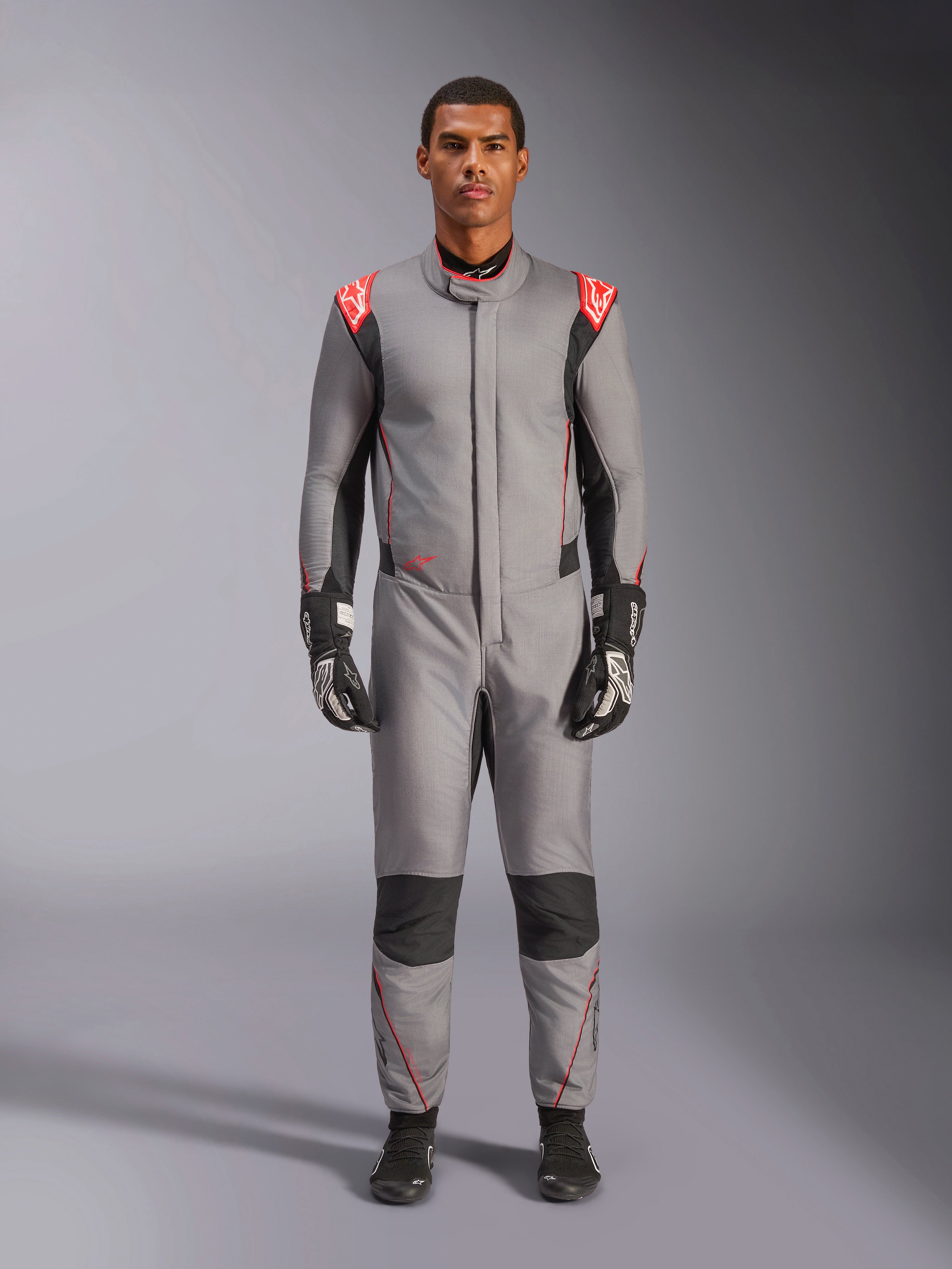 Alpinestars Hypertech V3 Suit, tuta da corsa professionale in grigio medio, nero e rosso fluo, indossata da un modello con guanti e scarpe da corsa neri, caratterizzata da una vestibilità atletica, accenti rosso fluo sulle spalle e pannelli neri su ginocchia e fianchi.