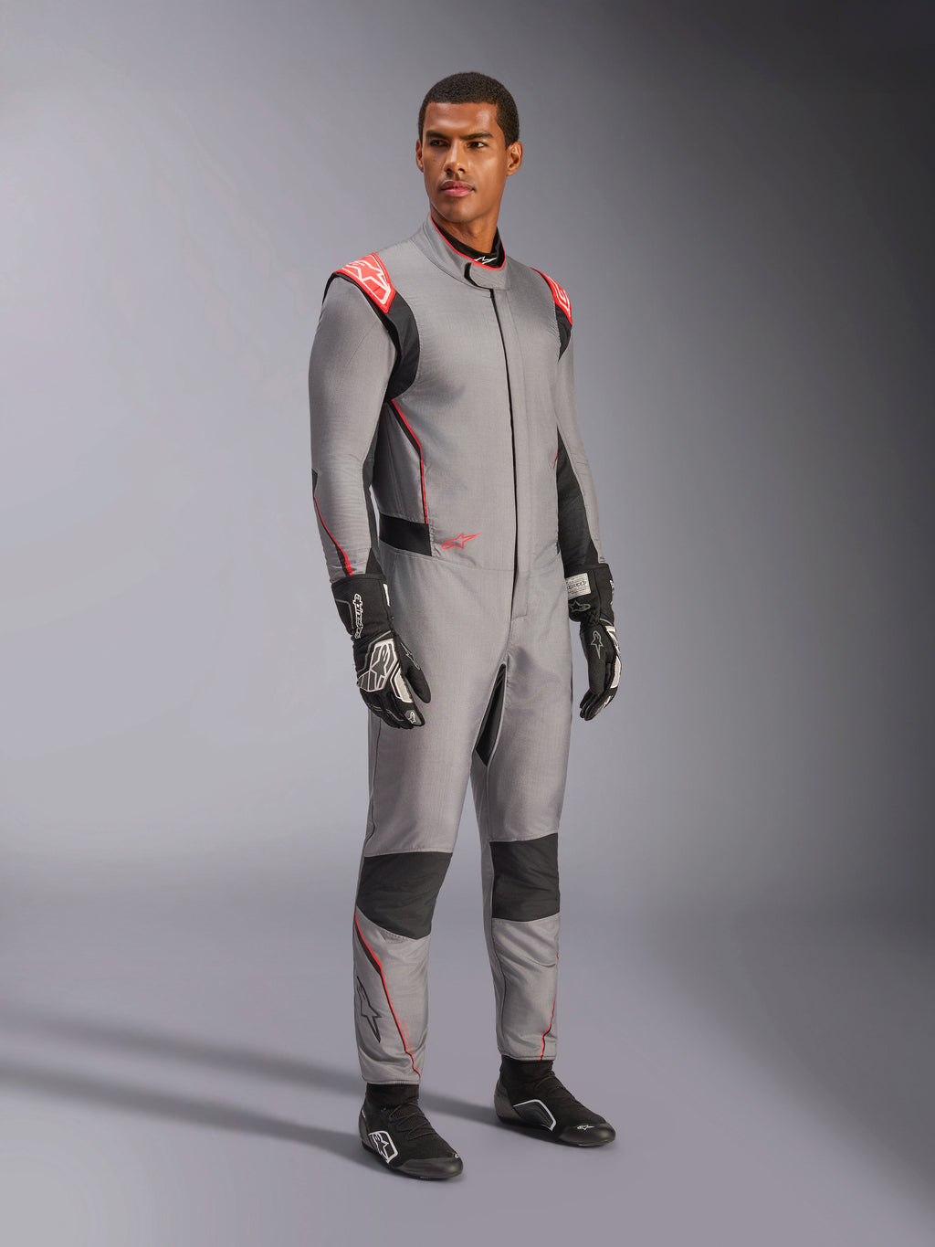 Modello che indossa la tuta Alpinestars Hypertech V3 in grigio medio con accenti neri e rosso fluo, tuta da corsa professionale abbinata a guanti e scarpe da racing neri, con struttura leggera in Nomex a due strati, dettagli rossi sulle spalle e vestibilità atletica