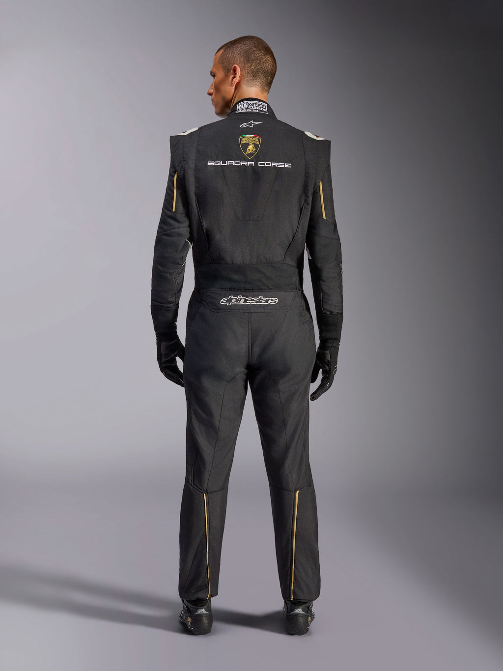 Alpinestars Edizione Limitata Lamborghini GP Tech V4 Suit FIA, tuta da corsa automobilistica, nera con accenti oro e argento, vista posteriore con marchio Lamborghini Squadra Corse, logo Alpinestars e bordini dorati su maniche e gambe, abbigliamento tecnico professionale