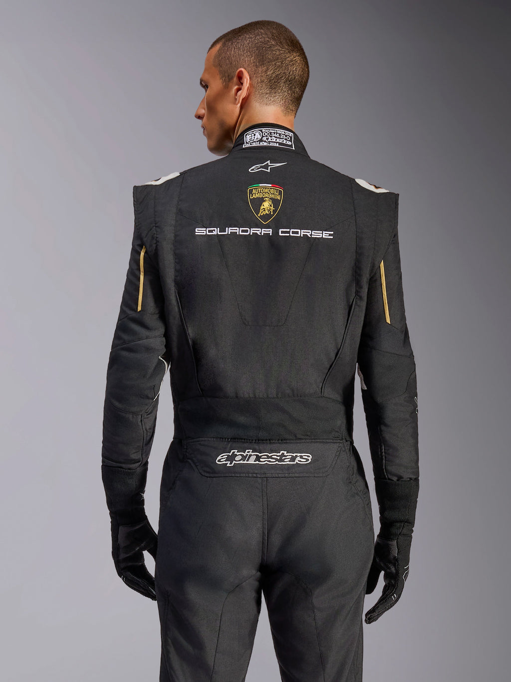 Vista posteriore della tuta Alpinestars Limited Edition Lamborghini GP Tech V4, nero con accenti oro, con scudetto Automobili Lamborghini e testo 'SQUADRA CORSE' sul retro, tuta da corsa professionale certificata FIA per competizioni automobilistiche