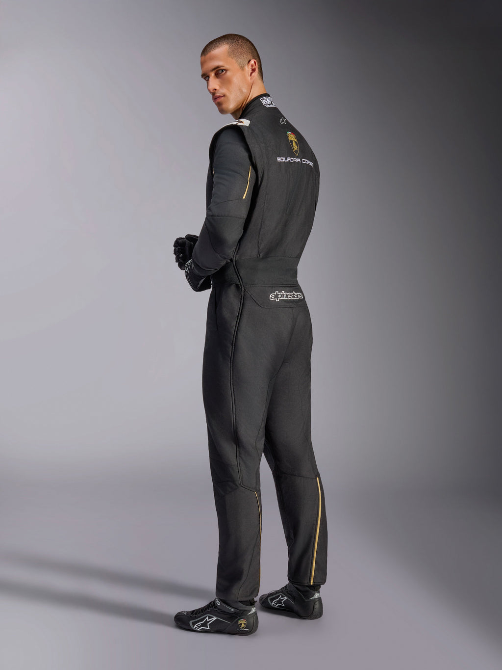 Tuta Alpinestars Limited Edition Lamborghini GP Tech V4, tuta da corsa professionale, nera con accenti argento e oro, vista posteriore che mostra il marchio Lamborghini Squadra Corse e Alpinestars, indossata con guanti e calzature da corsa neri, design motorsport raffinato