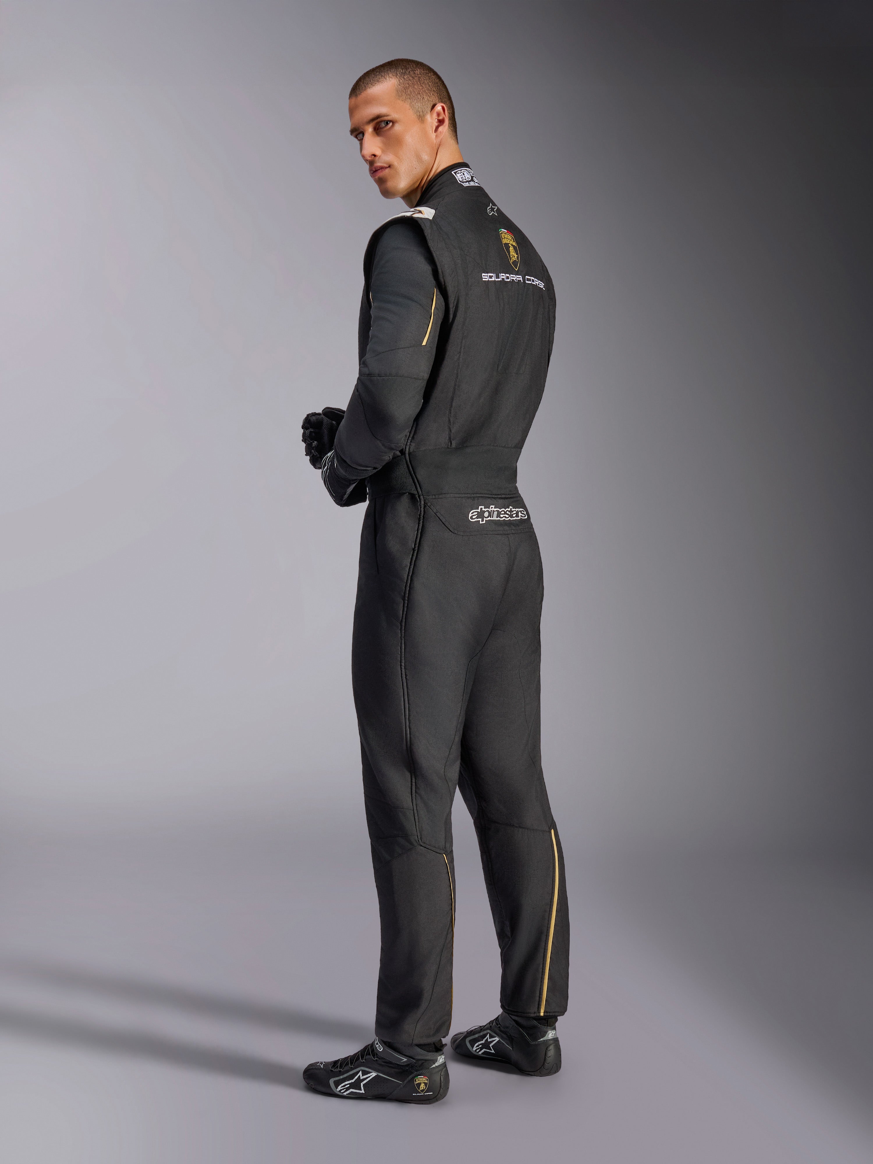 Tuta Alpinestars Limited Edition Lamborghini GP Tech V4, tuta da corsa professionale, nera con accenti argento e oro, vista posteriore che mostra il marchio Lamborghini Squadra Corse e Alpinestars, indossata con guanti e calzature da corsa neri, design motorsport raffinato