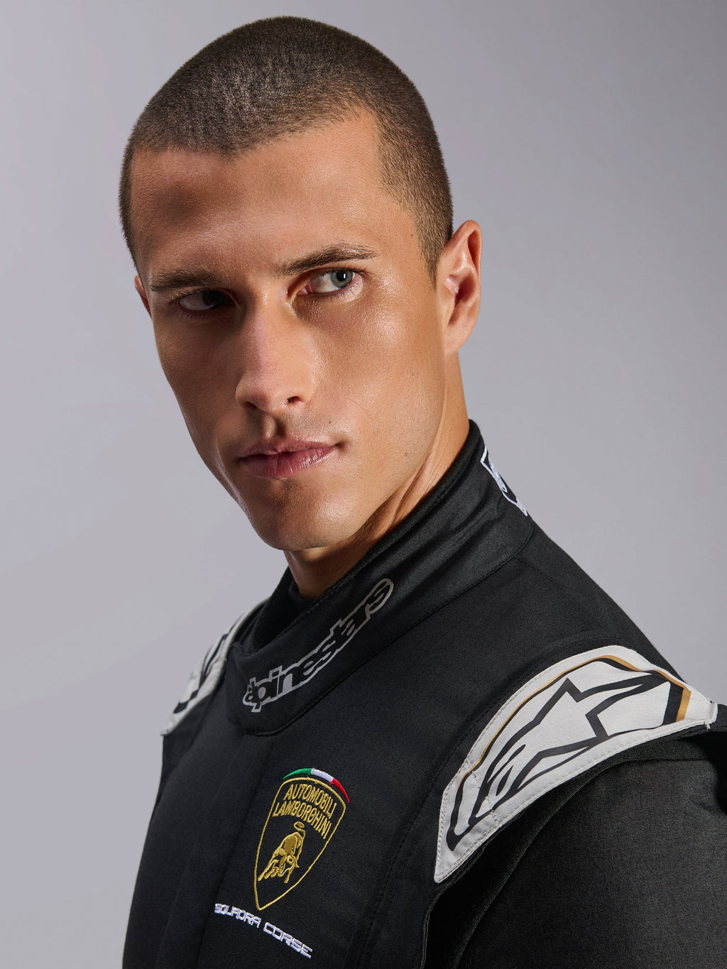 Primo piano di un modello che indossa la tuta Limited Edition Lamborghini GP Tech V4, nero, argento e oro, con loghi Automobili Lamborghini Squadra Corse e Alpinestars, abbigliamento professionale da corsa certificato FIA