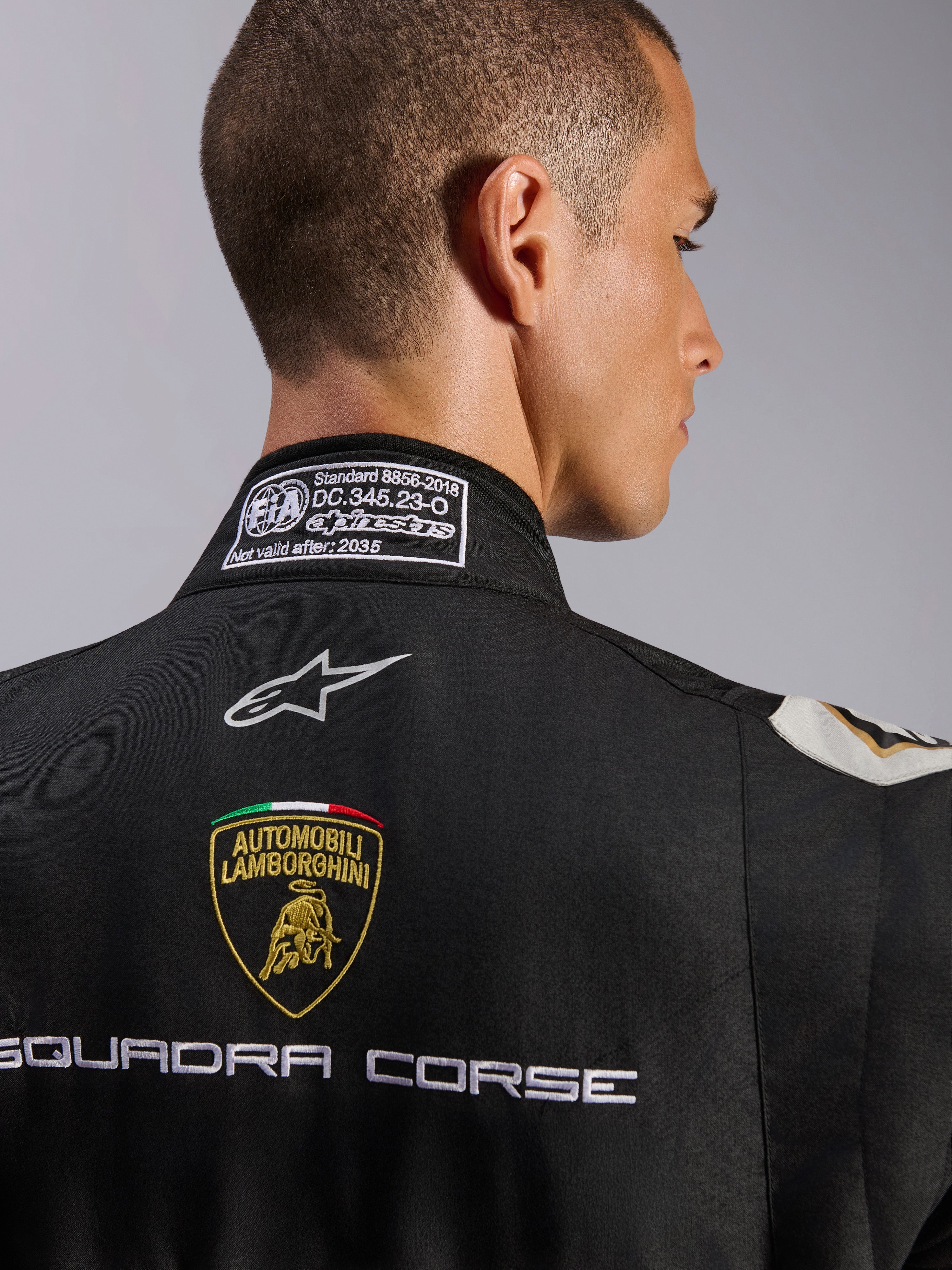 Alpinestars Limited Edition Lamborghini GP Tech V4 Tuta FIA, primo piano della parte superiore della schiena e del colletto, tuta nera con scudo Automobili Lamborghini e ricamo Squadra Corse, logo Alpinestars ed etichetta di certificazione FIA 8856-2018 sulla scollatura.