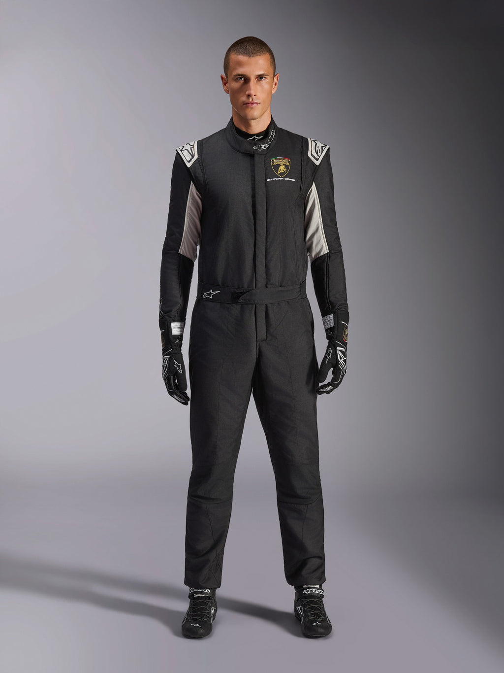 Tuta Alpinestars Limited Edition Lamborghini GP Tech V4 FIA, tuta da corsa automobilistica professionale, nera con accenti argento e oro, con logo Automobili Lamborghini Squadra Corse sul petto, indossata con guanti e stivali da corsa coordinati, design atletico ed elegante per competizioni in circuito