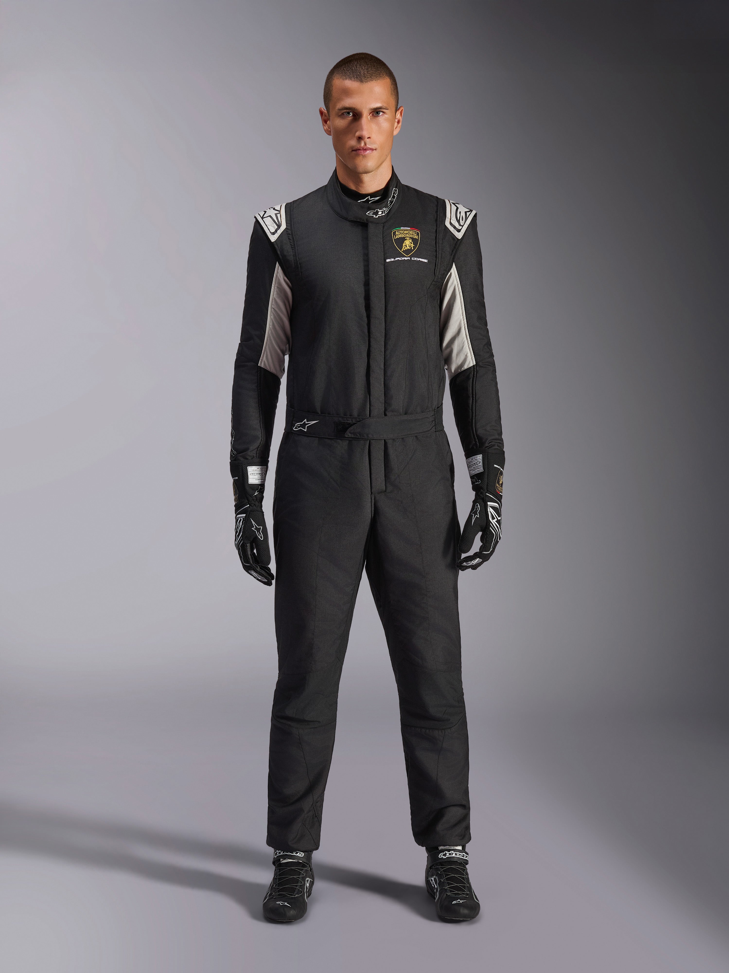 Tuta Alpinestars Limited Edition Lamborghini GP Tech V4 FIA, tuta da corsa automobilistica professionale, nera con accenti argento e oro, con logo Automobili Lamborghini Squadra Corse sul petto, indossata con guanti e stivali da corsa coordinati, design atletico ed elegante per competizioni in circuito