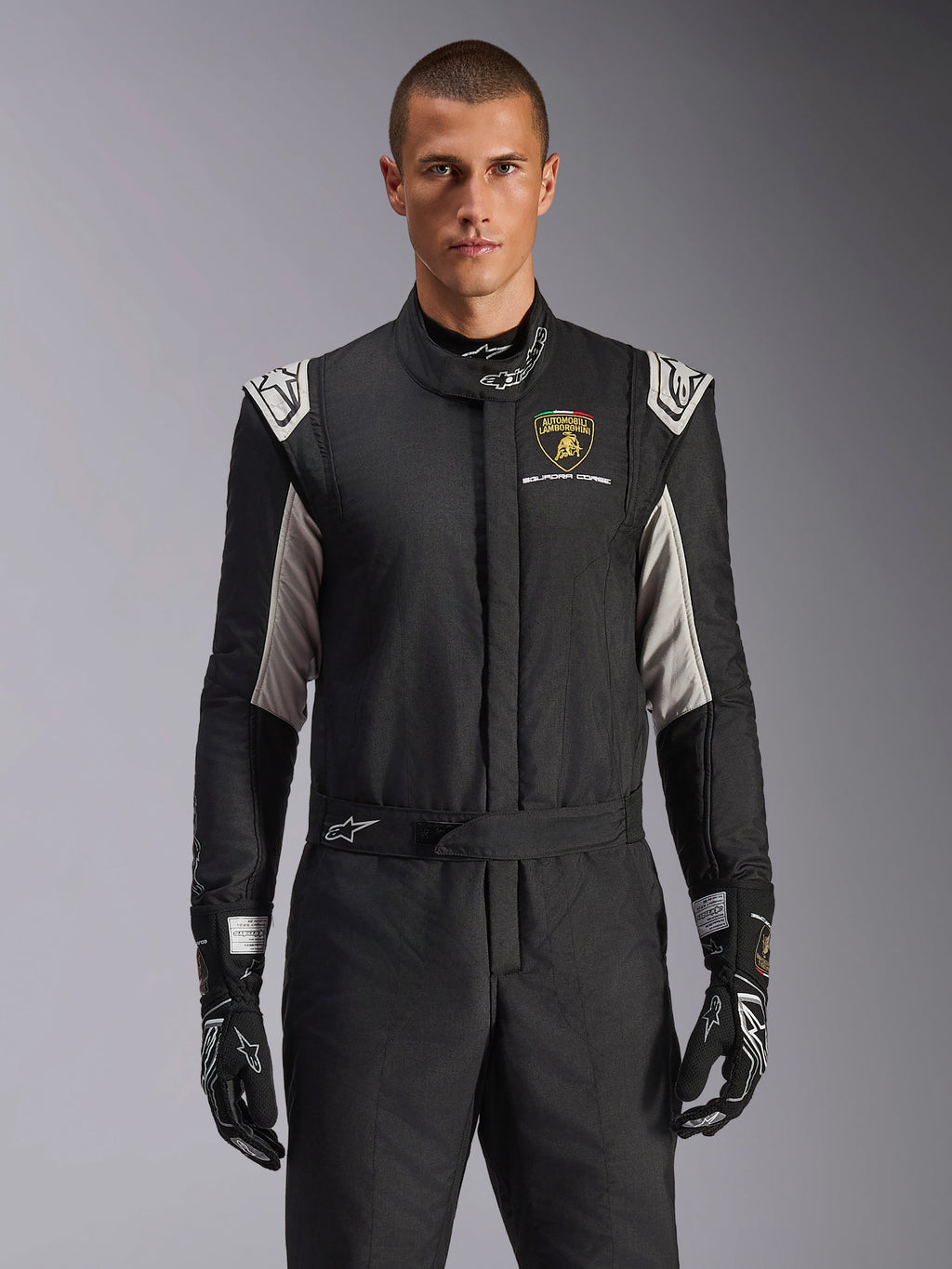 Alpinestars Limited Edition Lamborghini GP Tech V4 Suit, tuta da corsa professionale in nero con accenti argento e oro, con lo scudo Automobili Lamborghini Squadra Corse sul petto, indossata da un modello con guanti da corsa neri coordinati