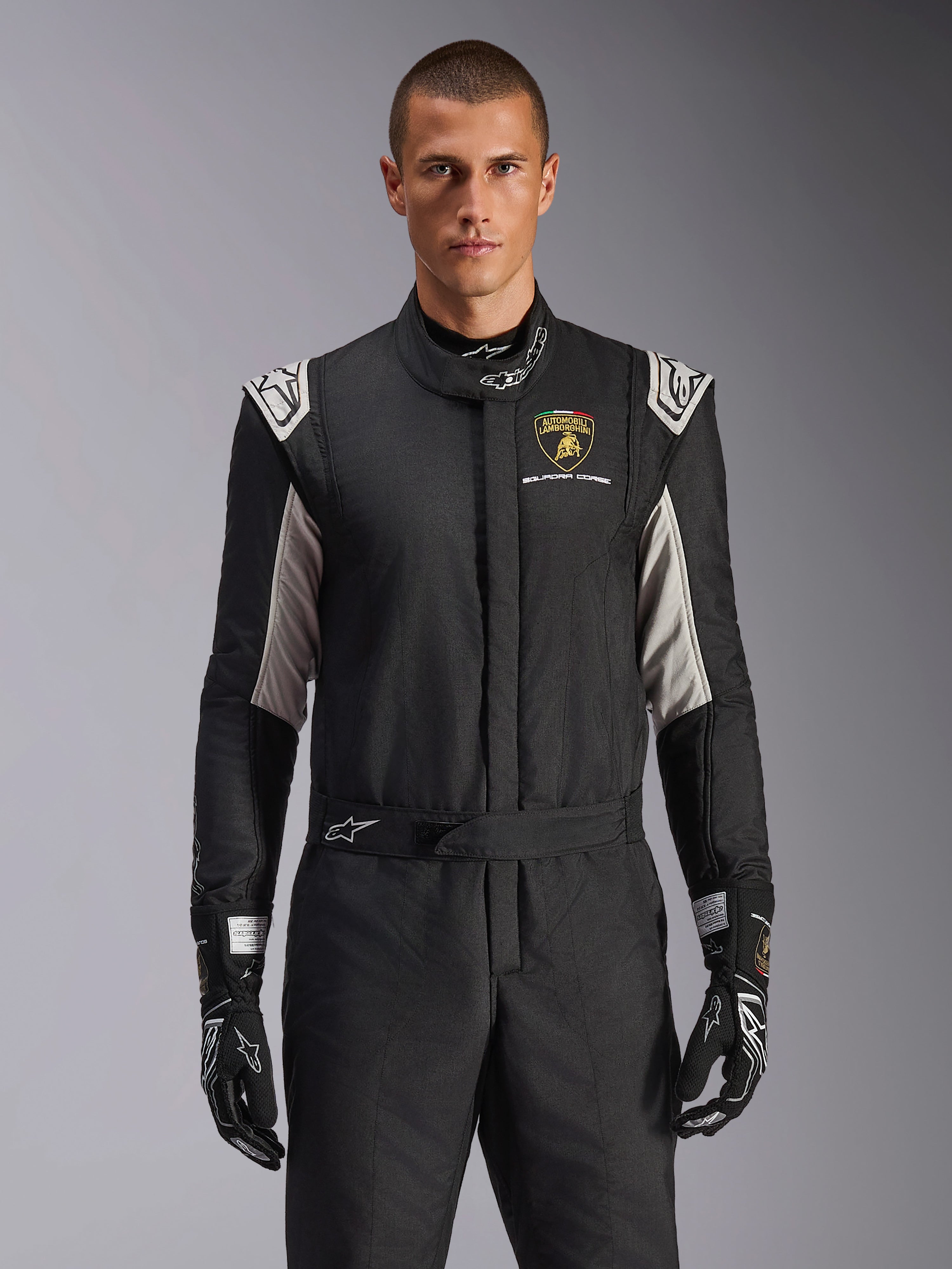 Alpinestars Limited Edition Lamborghini GP Tech V4 Suit, tuta da corsa professionale in nero con accenti argento e oro, con lo scudo Automobili Lamborghini Squadra Corse sul petto, indossata da un modello con guanti da corsa neri coordinati