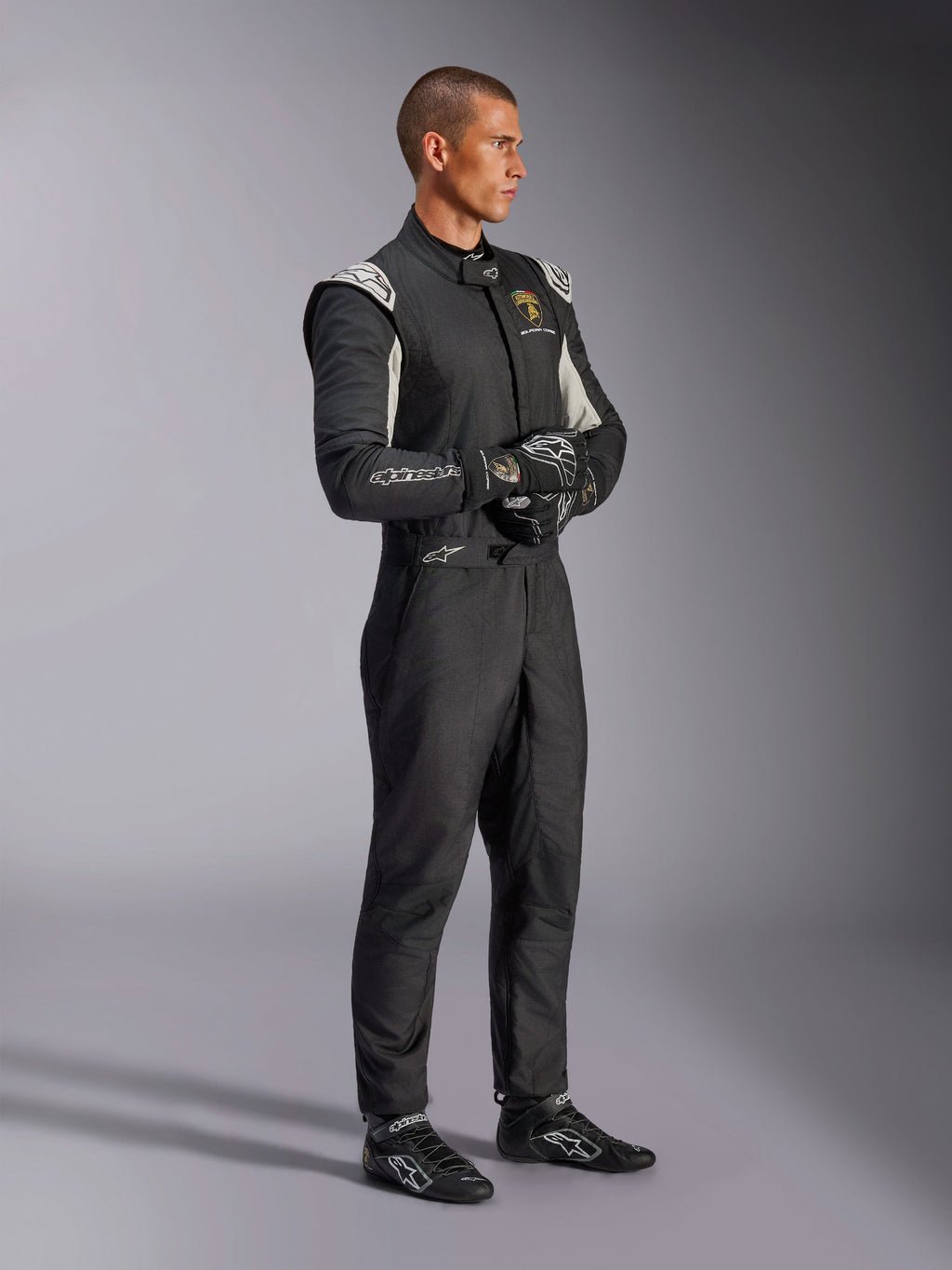 Pilota che indossa la tuta Alpinestars Limited Edition Lamborghini GP Tech V4, tuta da corsa certificata FIA in nero, argento e oro, abbinata a guanti e scarpe coordinate, con marchio Lamborghini Squadra Corse, abbigliamento professionale per motorsport