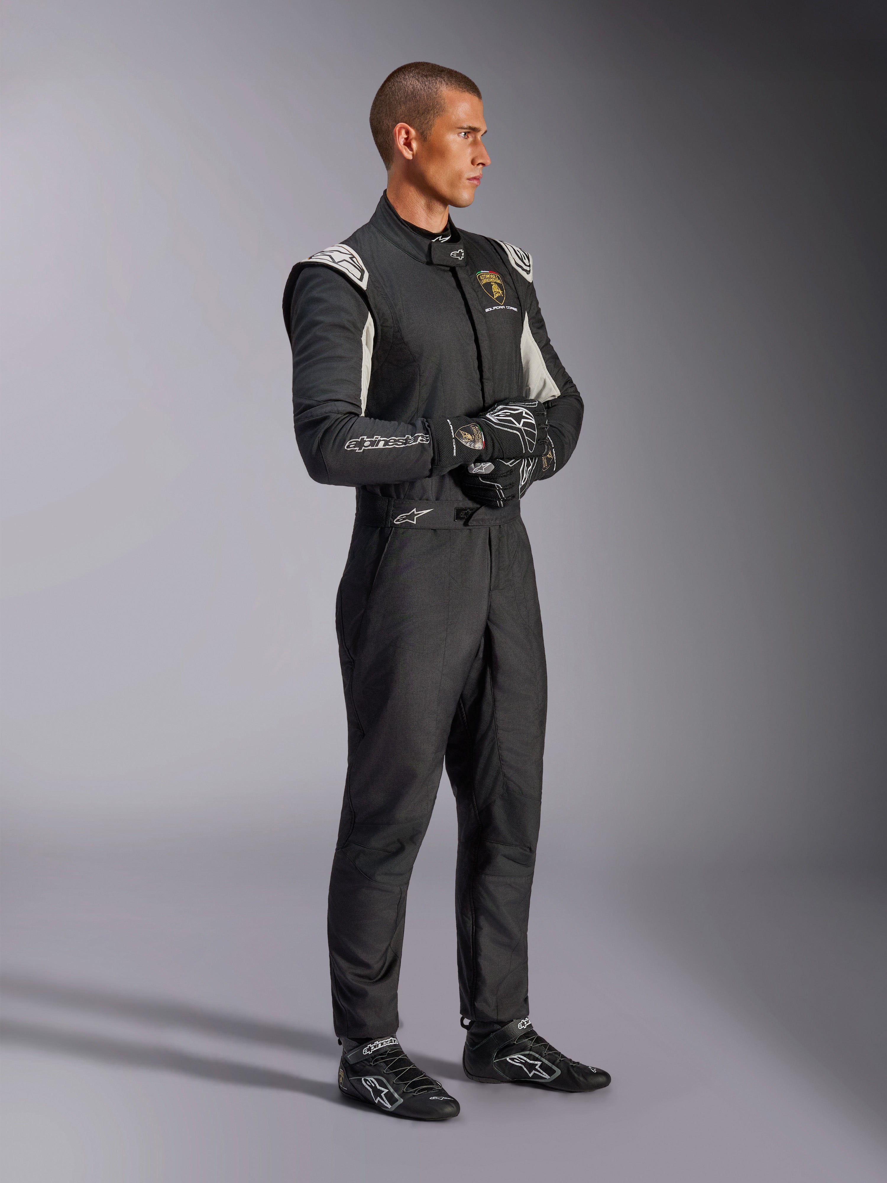 Pilota che indossa la tuta Alpinestars Limited Edition Lamborghini GP Tech V4, tuta da corsa certificata FIA in nero, argento e oro, abbinata a guanti e scarpe coordinate, con marchio Lamborghini Squadra Corse, abbigliamento professionale per motorsport