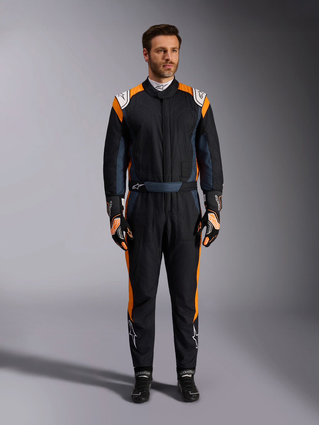 Alpinestars GP Pro Comp V2 FIA Suit, tuta da auto racing nero asfalto e arancione fluo, modello uomo che indossa equipaggiamento protettivo completo con guanti e scarpe da corsa abbinati, caratterizzata da loghi Alpinestars bianchi e vivaci pannelli laterali arancioni, abbigliamento professionale per motorsport