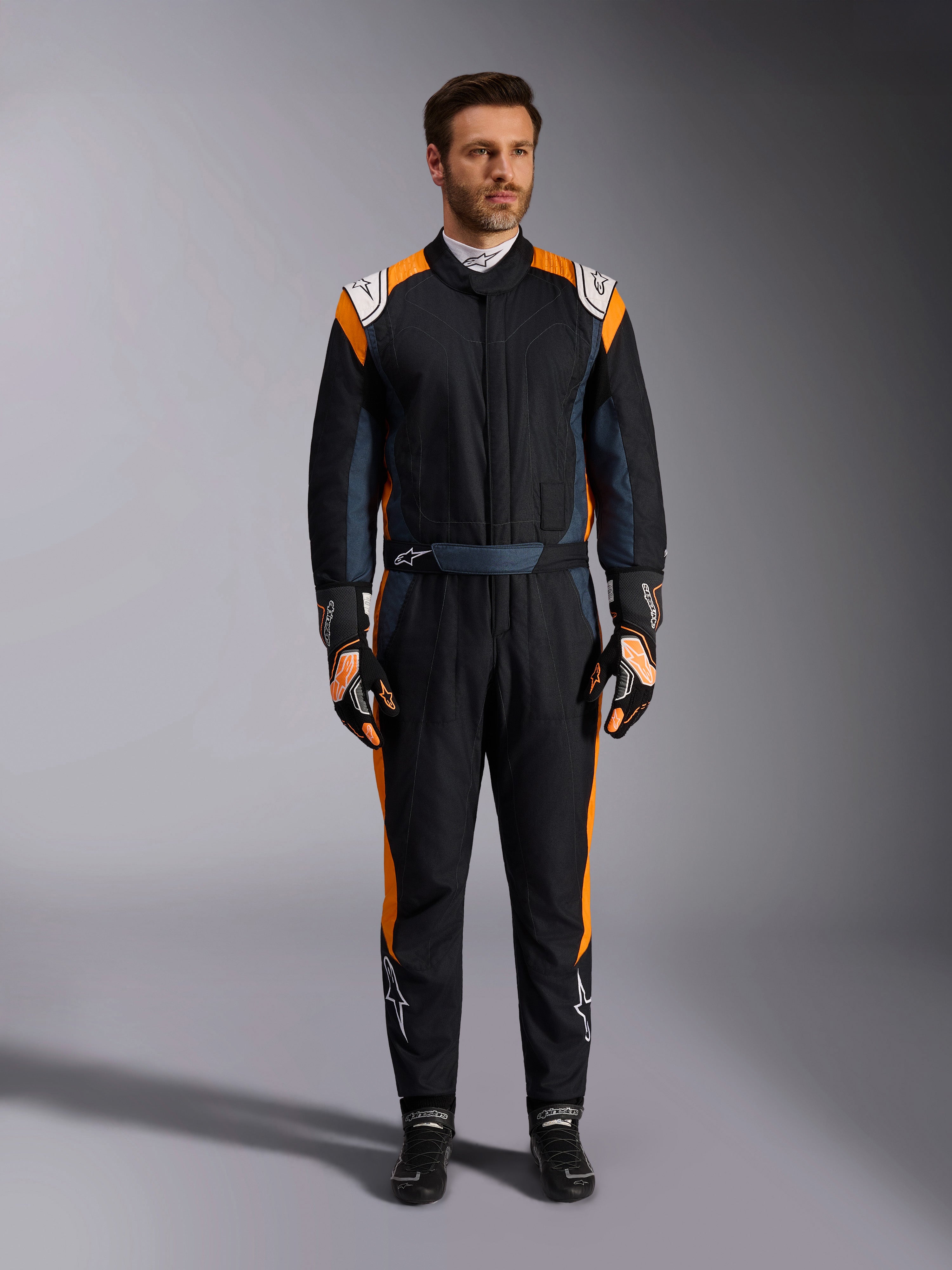 Alpinestars GP Pro Comp V2 FIA Suit, tuta da auto racing nero asfalto e arancione fluo, modello uomo che indossa equipaggiamento protettivo completo con guanti e scarpe da corsa abbinati, caratterizzata da loghi Alpinestars bianchi e vivaci pannelli laterali arancioni, abbigliamento professionale per motorsport