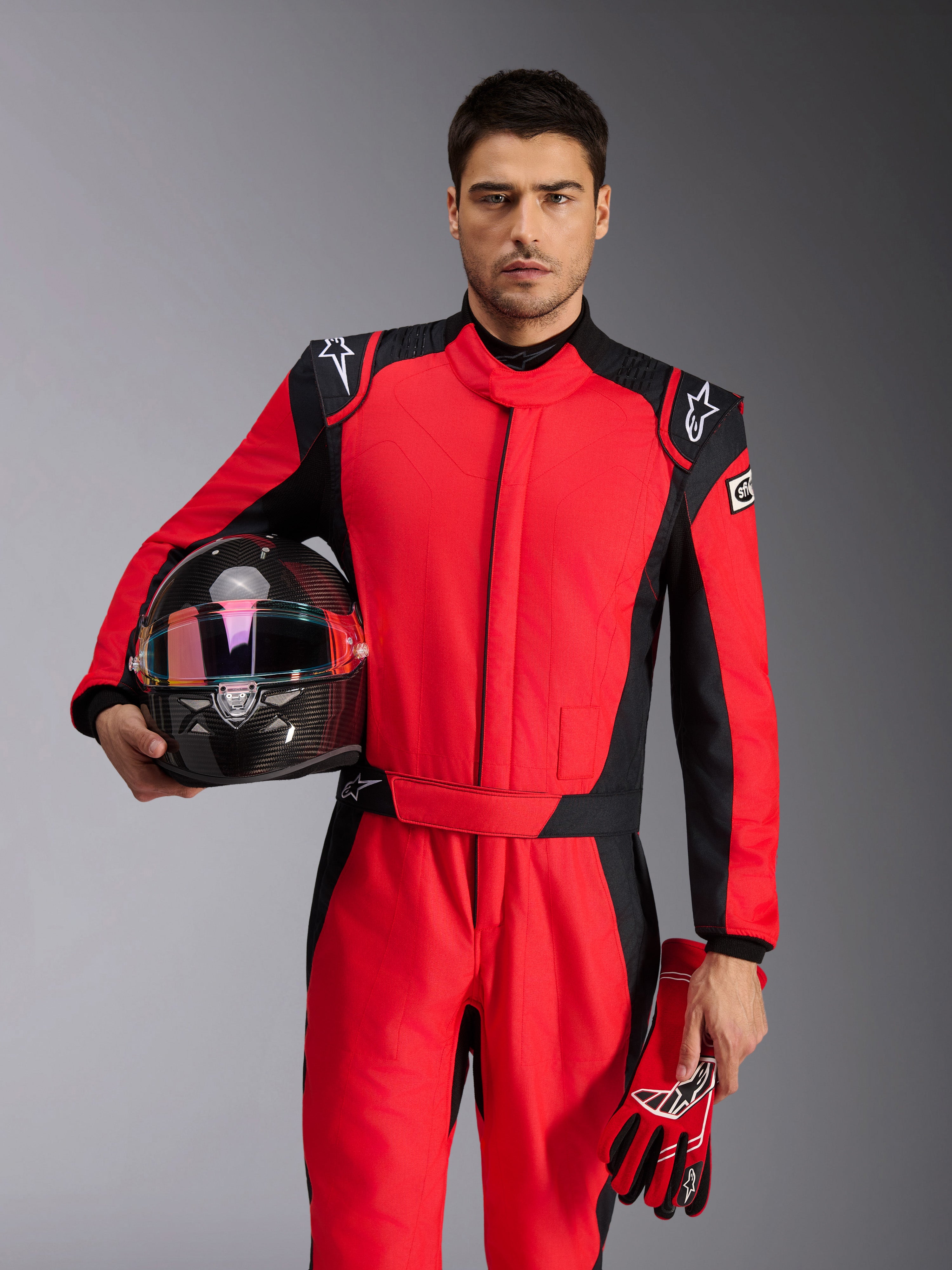 Pilota Alpinestars con tuta GP Pro Comp V2 FIA in rosso e nero, tiene un casco da corsa in fibra di carbonio e guanti coordinati, abbigliamento professionale da motorsport