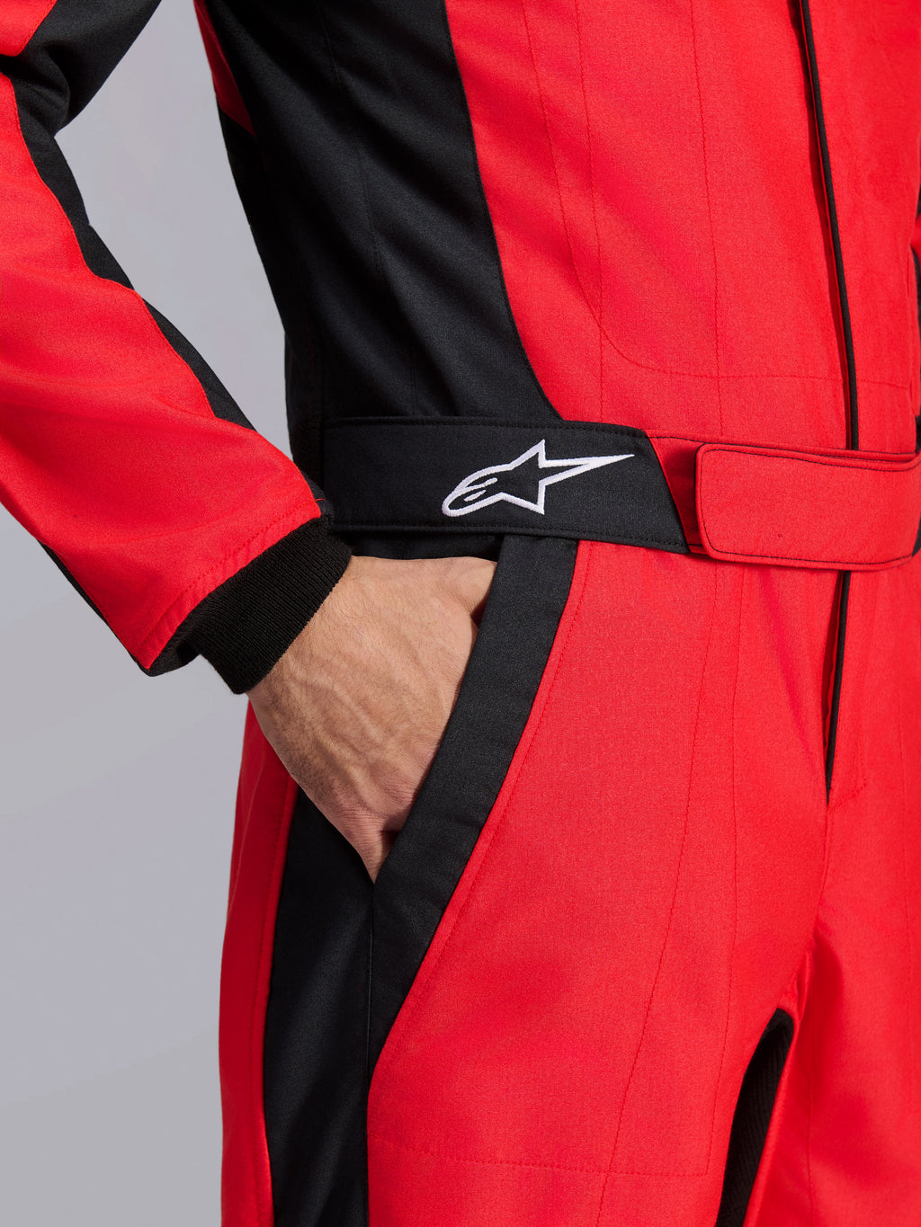 Tuta da gara Alpinestars GP Pro Comp V2 FIA, rosso e nero, primo piano della zona della vita con una mano in tasca, cintura nera con logo a stella bianco e pannelli strutturati per l'auto racing