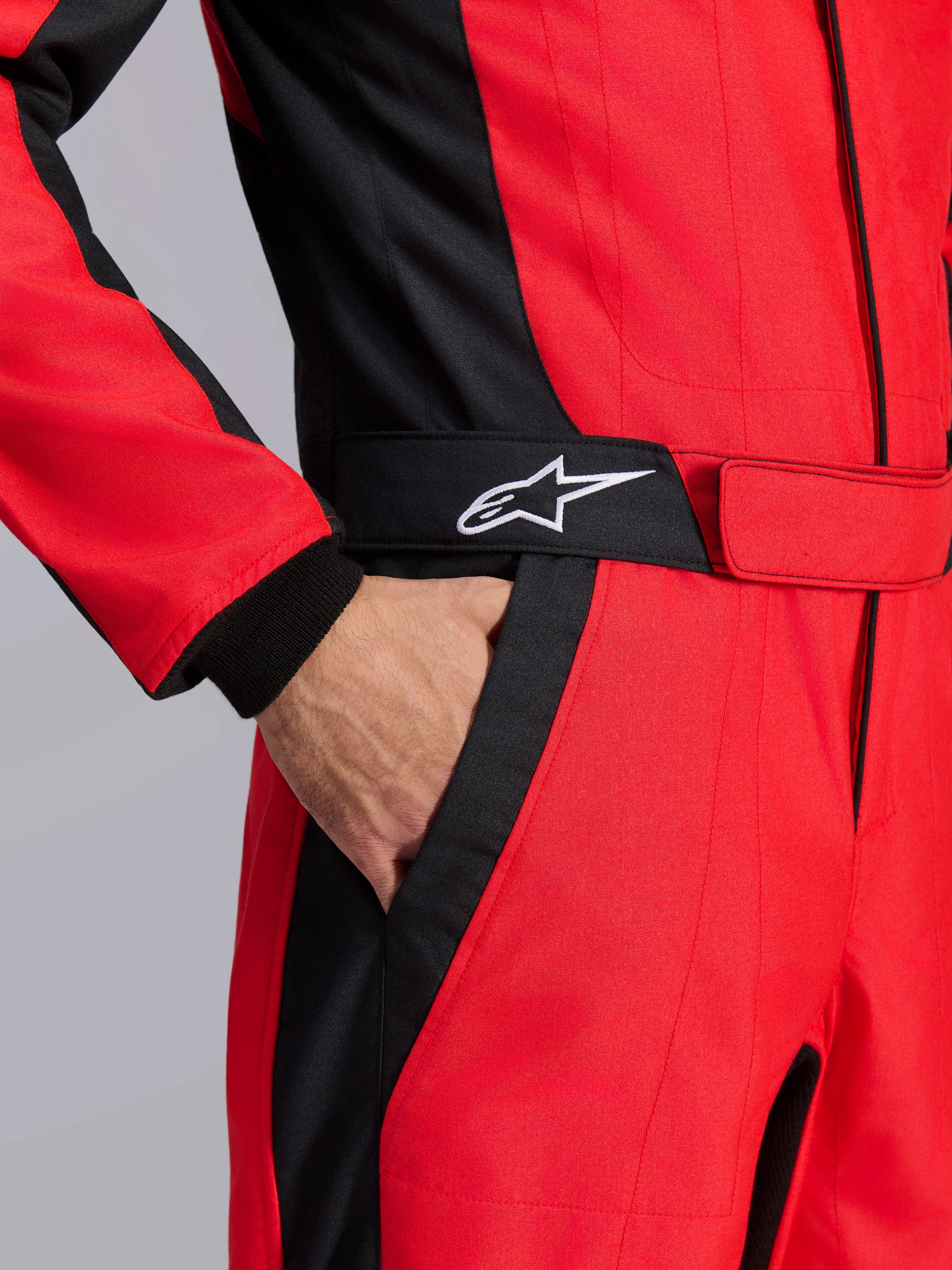 Tuta da gara Alpinestars GP Pro Comp V2 FIA, rosso e nero, primo piano della zona della vita con una mano in tasca, cintura nera con logo a stella bianco e pannelli strutturati per l'auto racing