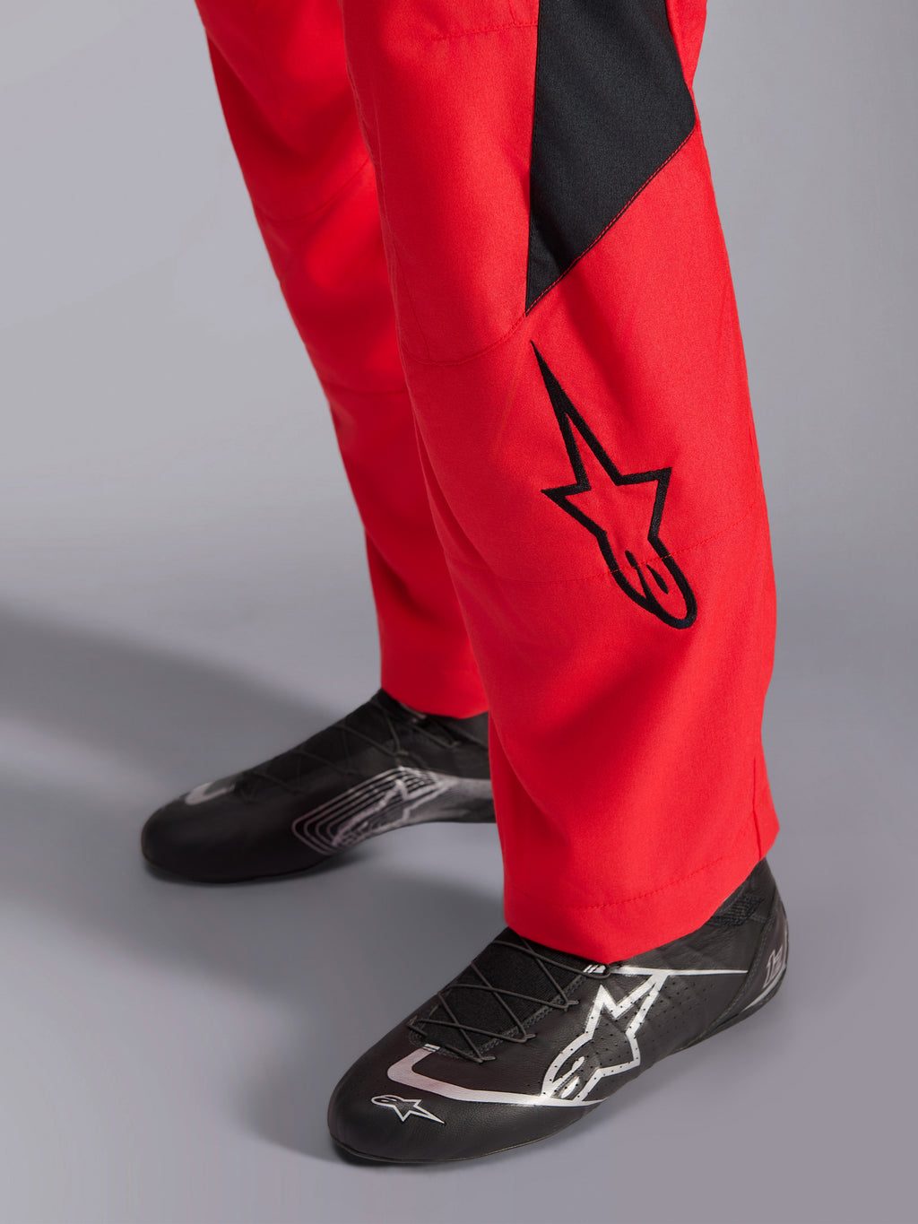 Tuta da gara Alpinestars GP Pro Comp V2 FIA, primo piano della parte inferiore della gamba e delle scarpe da corsa, rosso con pannello e logo Astars nero, abbinata a calzature tecniche nere, equipaggiamento per corse automobilistiche