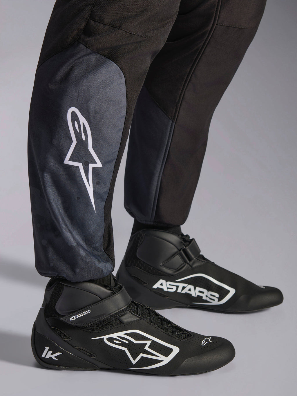 Alpinestars Tuta KMX-5 V3, Black Dark Shadow, primo piano della parte inferiore delle gambe e delle caviglie, logo a stella bianco sul polpaccio, abbinata a scarpe da kart nere, con dettagli sui polsini delle caviglie in maglia e vestibilità athletic-regular.