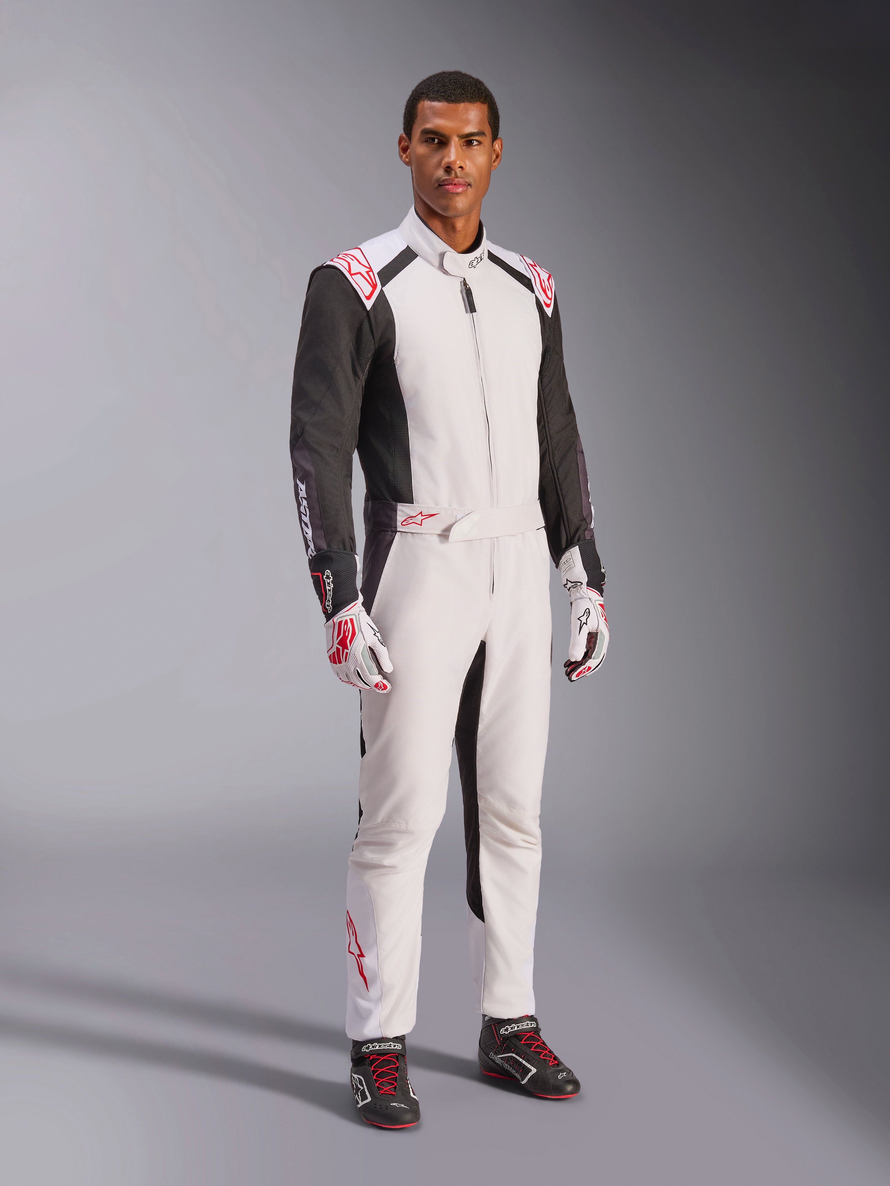 Tuta Alpinestars KMX-5 V3, bianco, nero e rosso brillante, tuta intera da karting indossata con guanti coordinati bianchi e rossi e scarpe da corsa nere, con maniche nere e vivaci accenti rossi, equipaggiamento professionale da motorsport per karting competitivo.