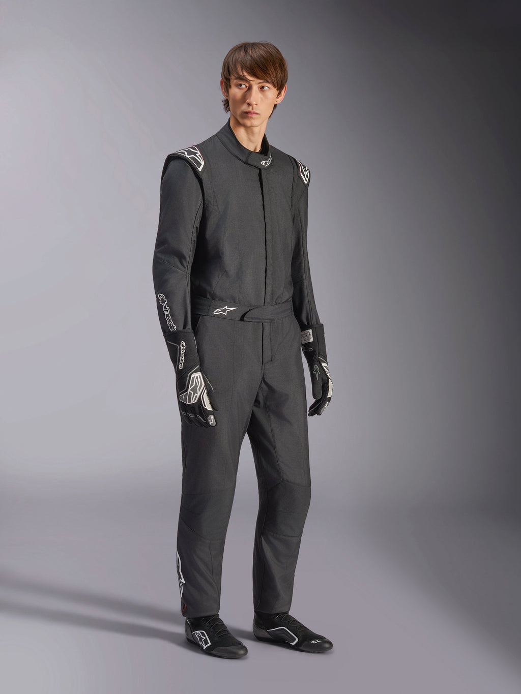 Tuta Alpinestars GP Tech V4, tuta auto intera, nero e bianco, indossata con guanti e calzature da corsa abbinati, caratterizzata da loghi bianchi su spalle e avambracci, design professionale per il motorsport